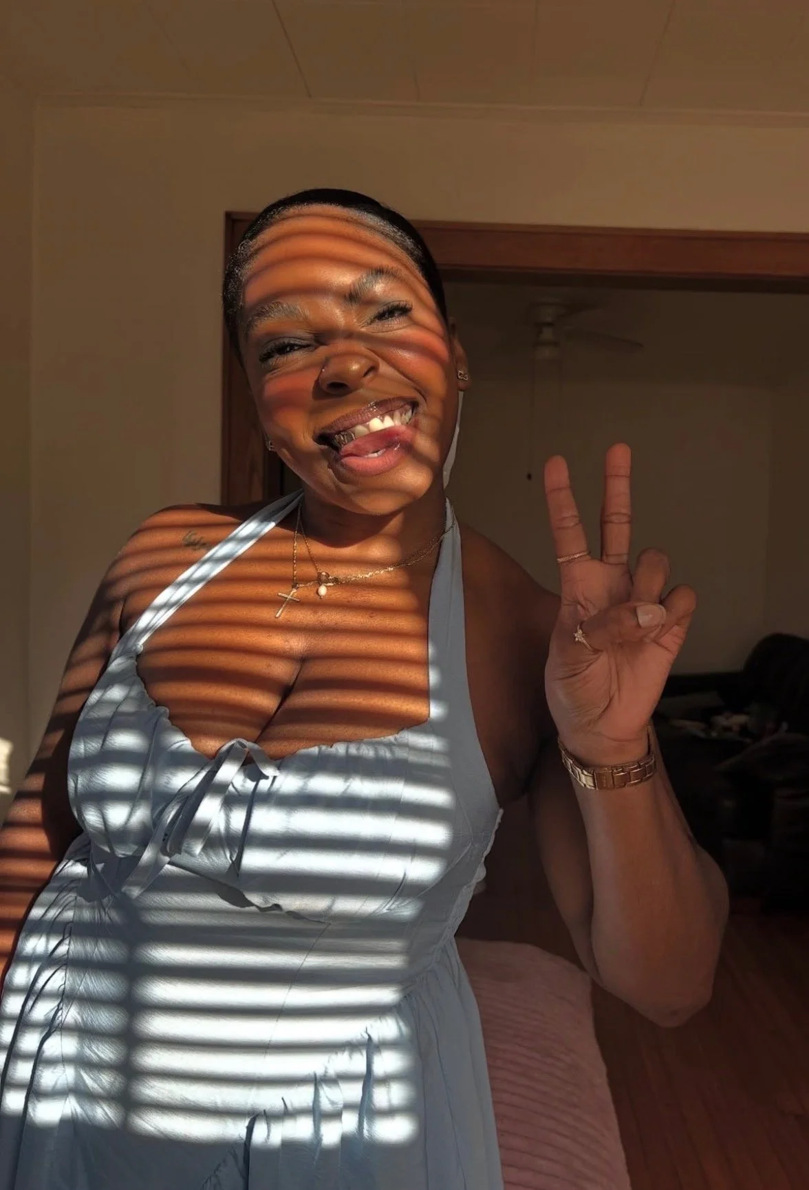A woman smiling and making a peace sign with her fingers, standing near a window with blinds that cast striped shadows on her face and body.