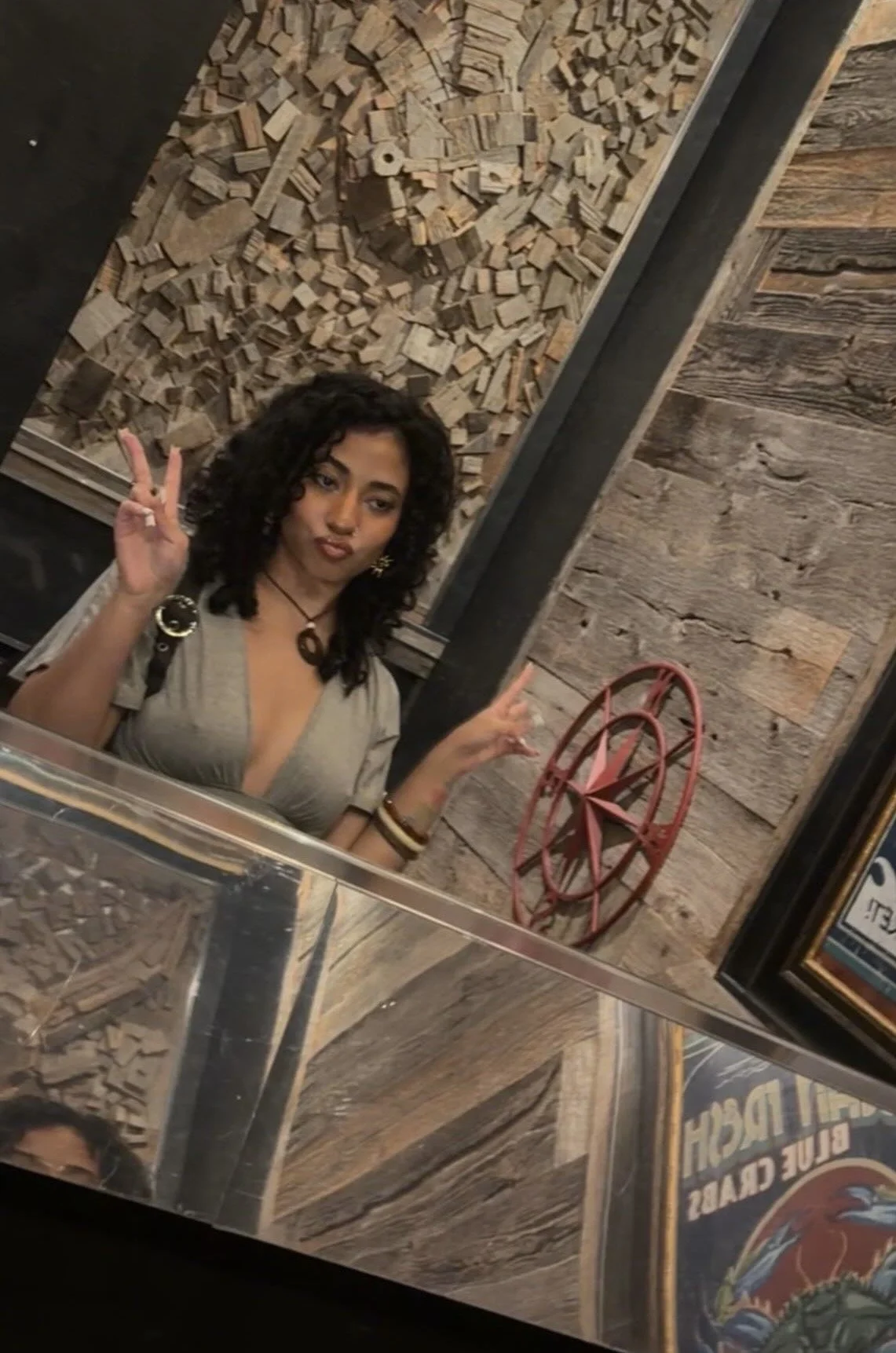 A woman with dark curly hair wearing layered necklaces and a sleeveless patterned top. She is sitting indoors against a wood-paneled background.