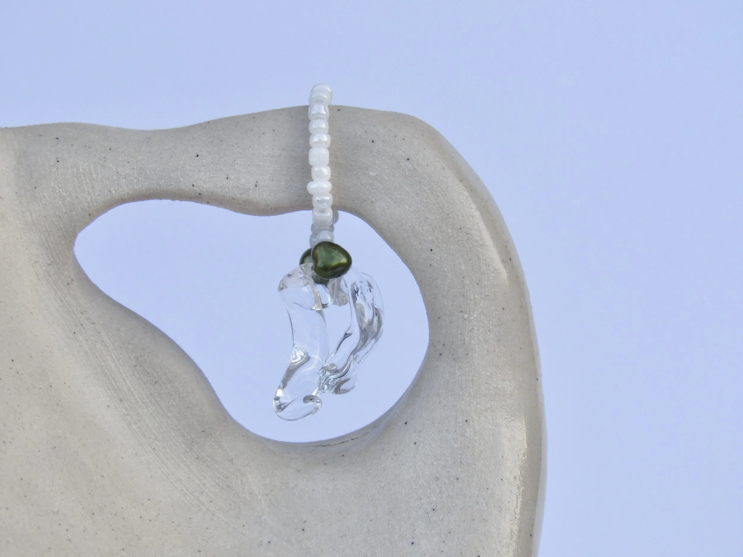 A piece of jewelry with white beads and a green stone, hanging on a light-colored, textured ceramic or stone sculpture with a circular opening, against a pale blue background.