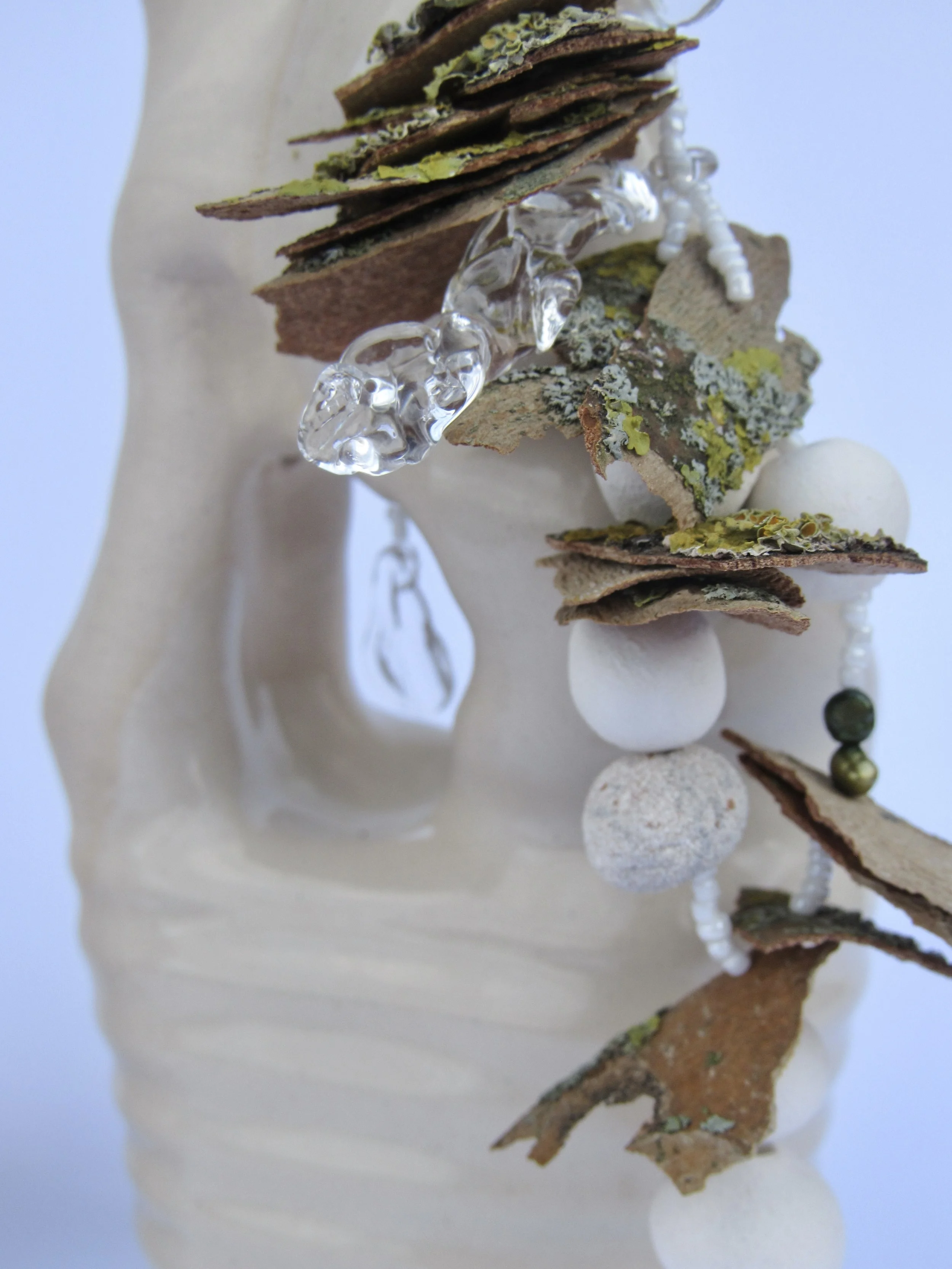 A close-up of an art piece featuring dried bark, lichen, stones, and beads arranged on a neutral-colored sculpture or mannequin.