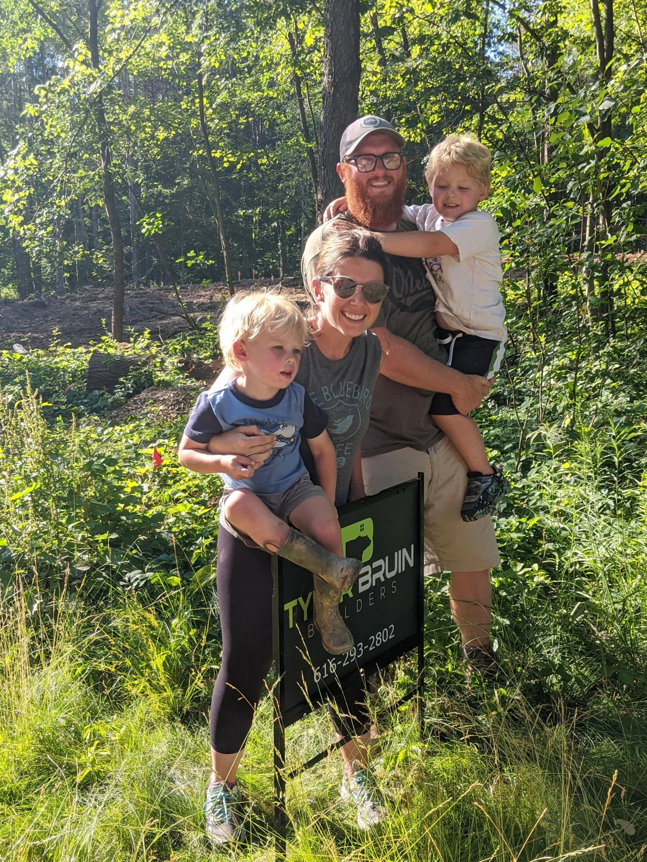 A family of four posing outdoors in a lush green forest. An adult woman is holding a young boy, and a man with a beard and glasses is carrying another boy on his shoulders. There is a black and green sign with contact information in front of them.