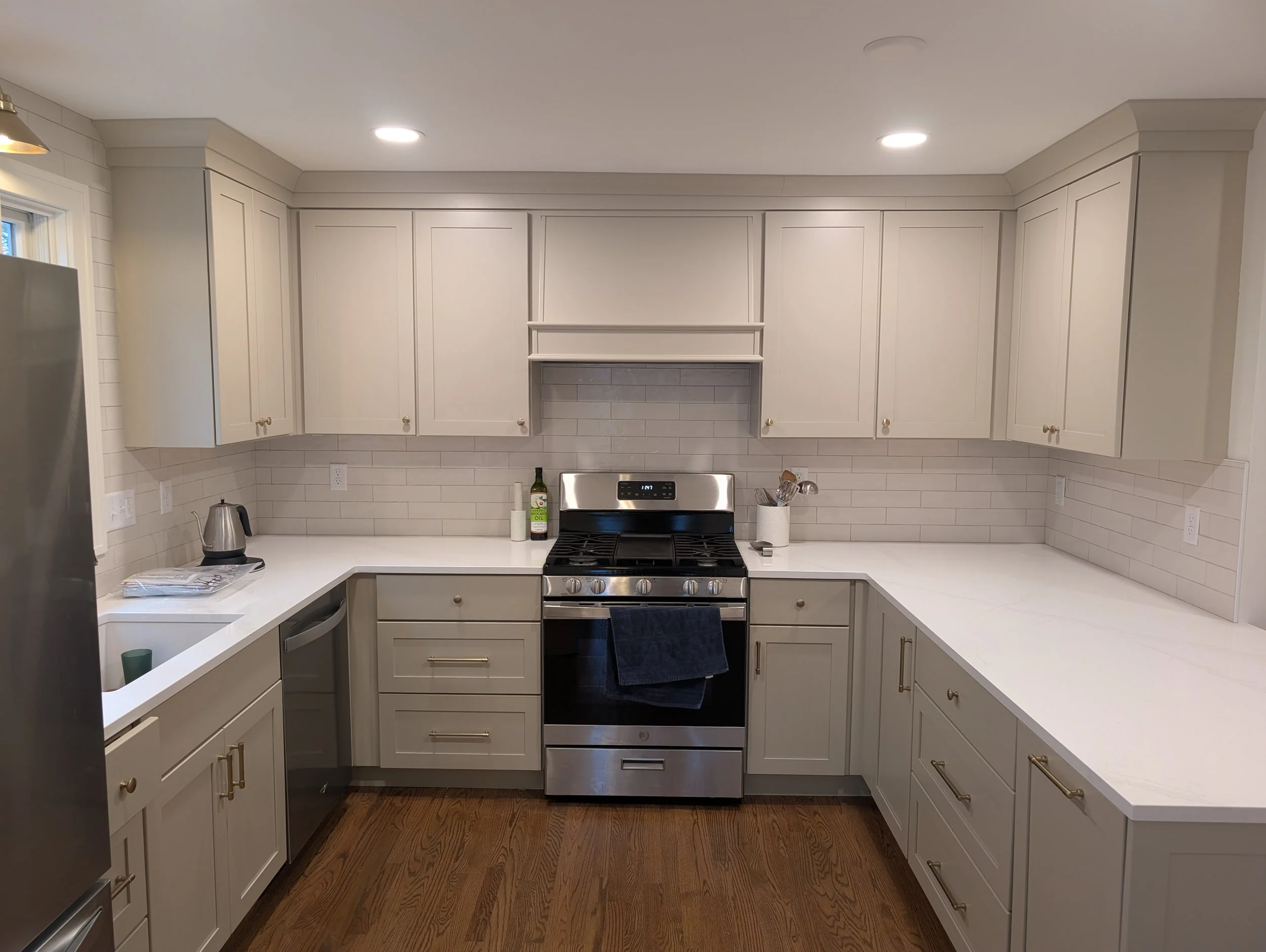 White kitchen cabinets above a stove with a gray backsplash, a bottle of cooking oil, a paper towel roll, and kitchen utensils on the counter.