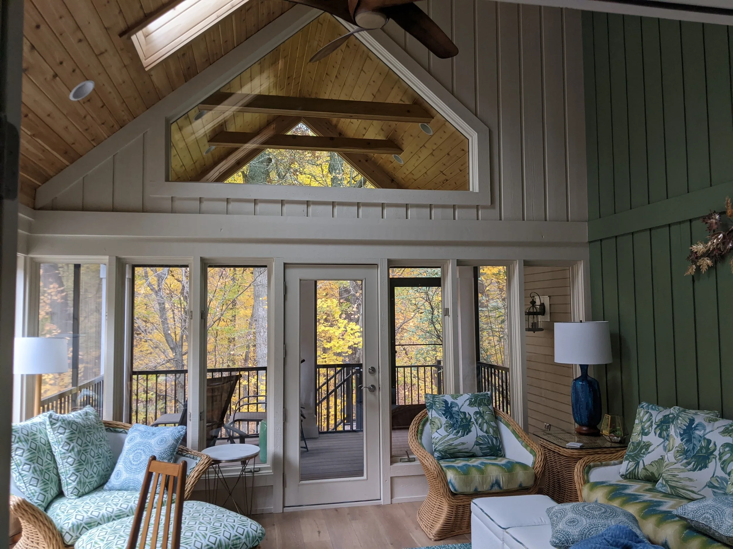 Living room with vaulted wooden ceiling, glass doors leading to a deck, and a view of trees with fall foliage outside.