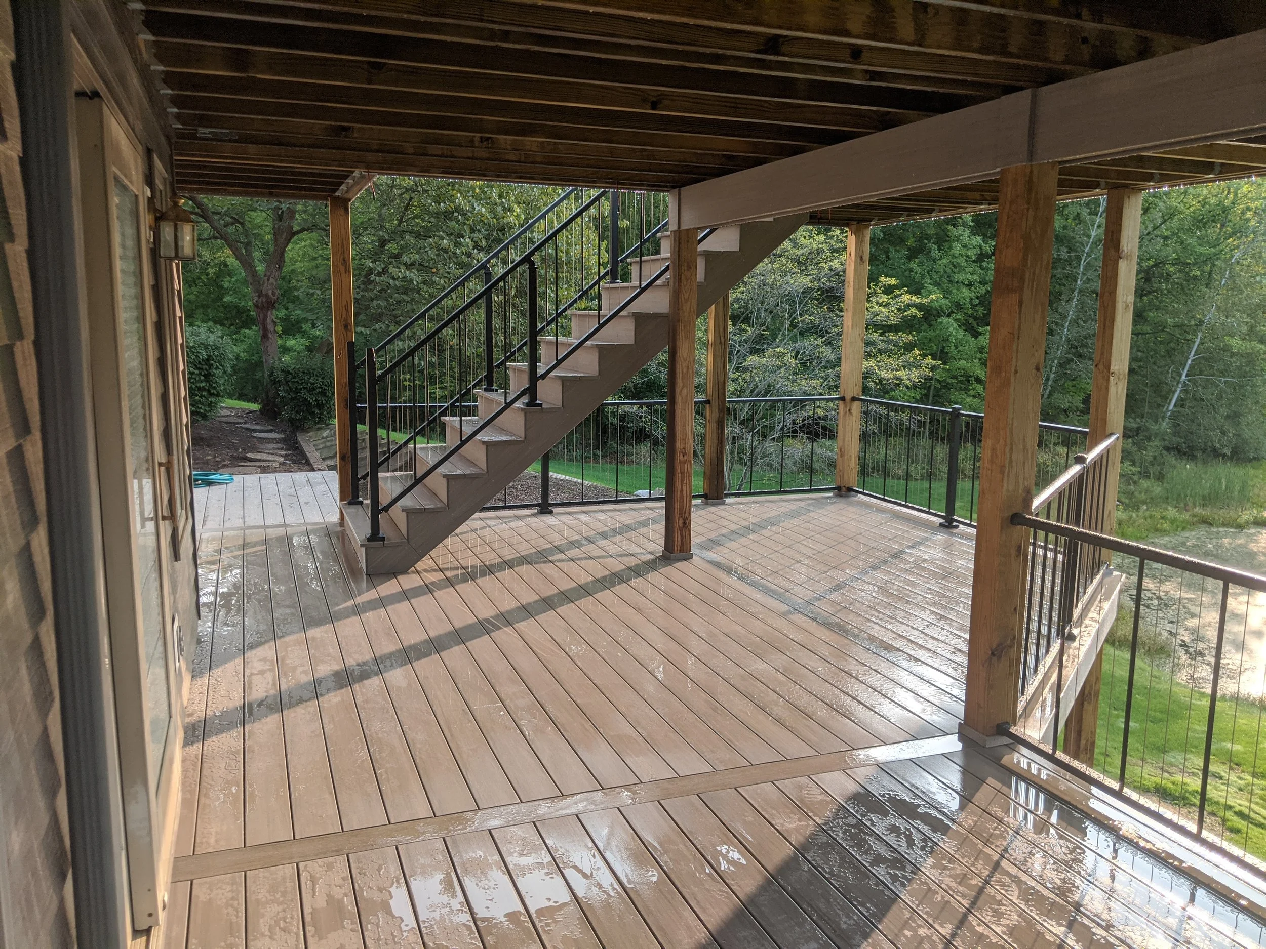 Wooden deck with stairs leading down, surrounded by trees and greenery, with some wet surface indicating recent rain.