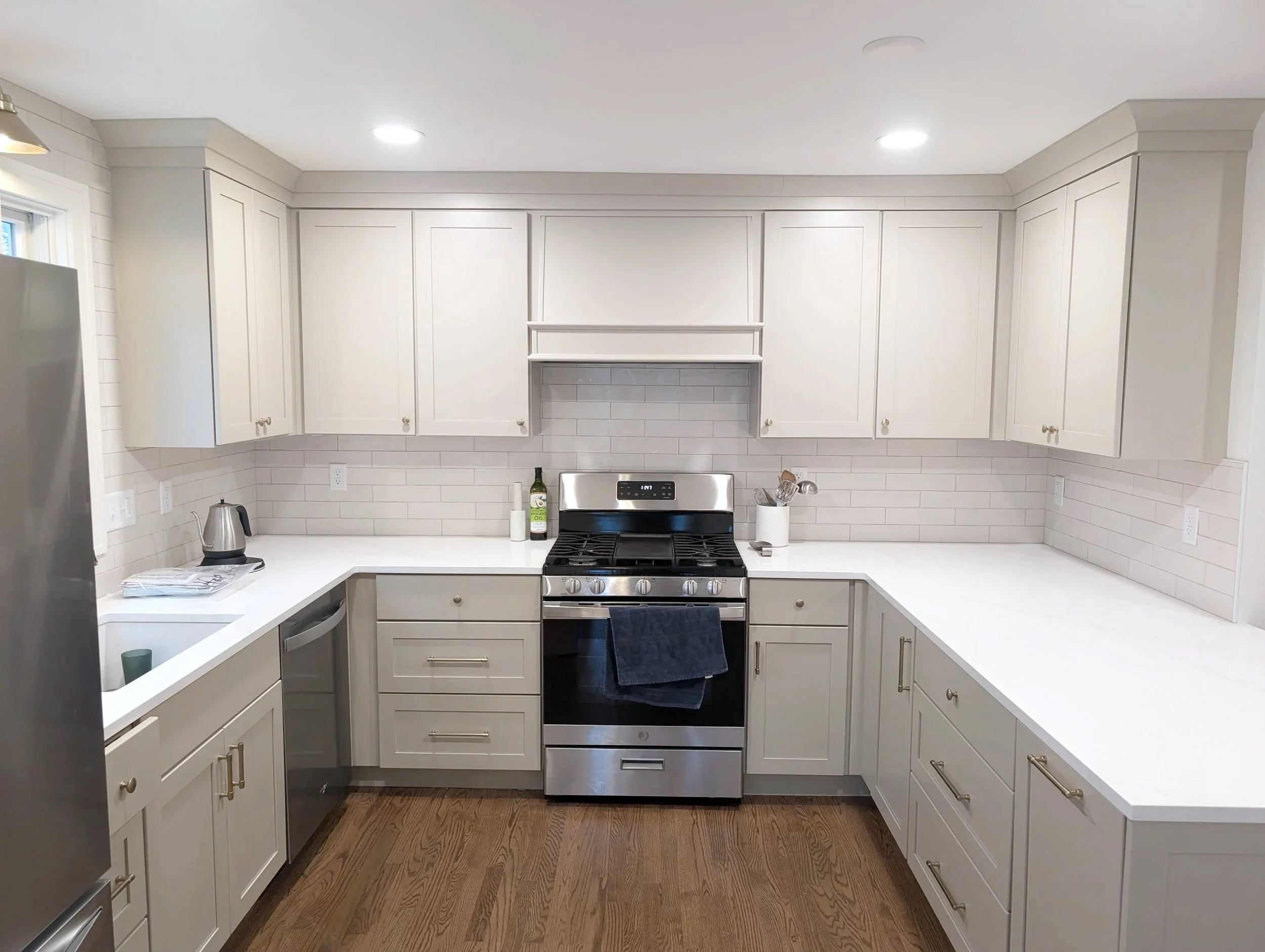 Modern kitchen with white cabinets, stainless steel appliances, wooden floor, and a white countertop. Items on the counter include a kettle, paper towels, and kitchen utensils.