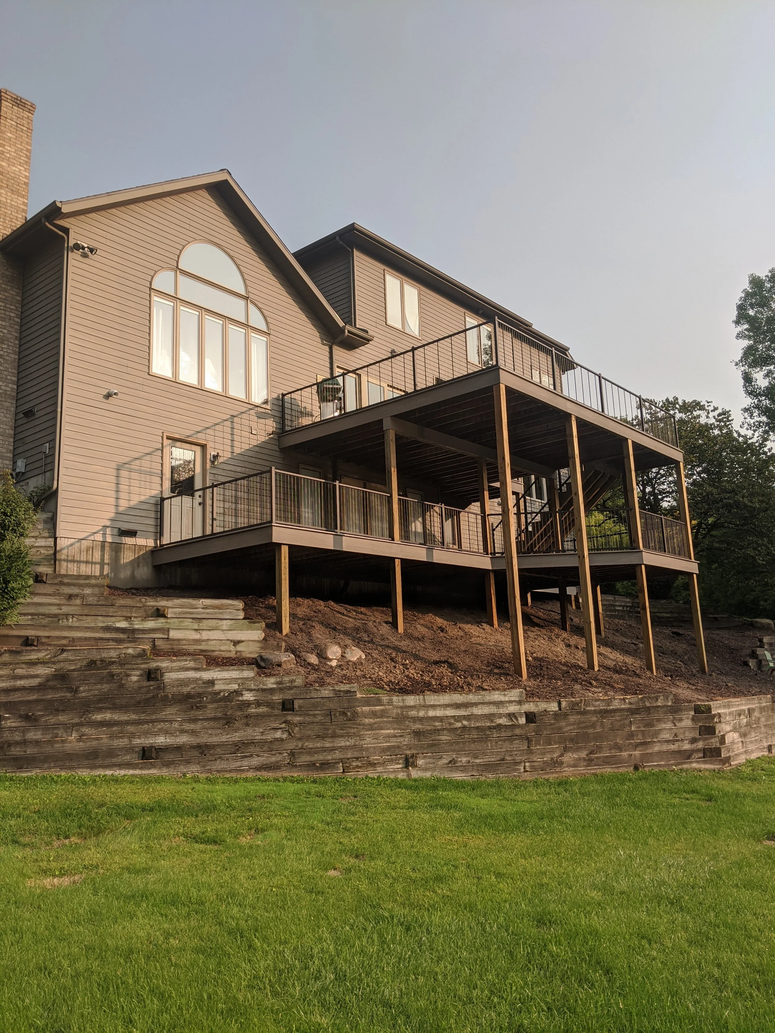A multi-story house with a light brown exterior, large arched windows, and multiple decks supported by wooden beams, set against a clear sky with lush greenery in the foreground.