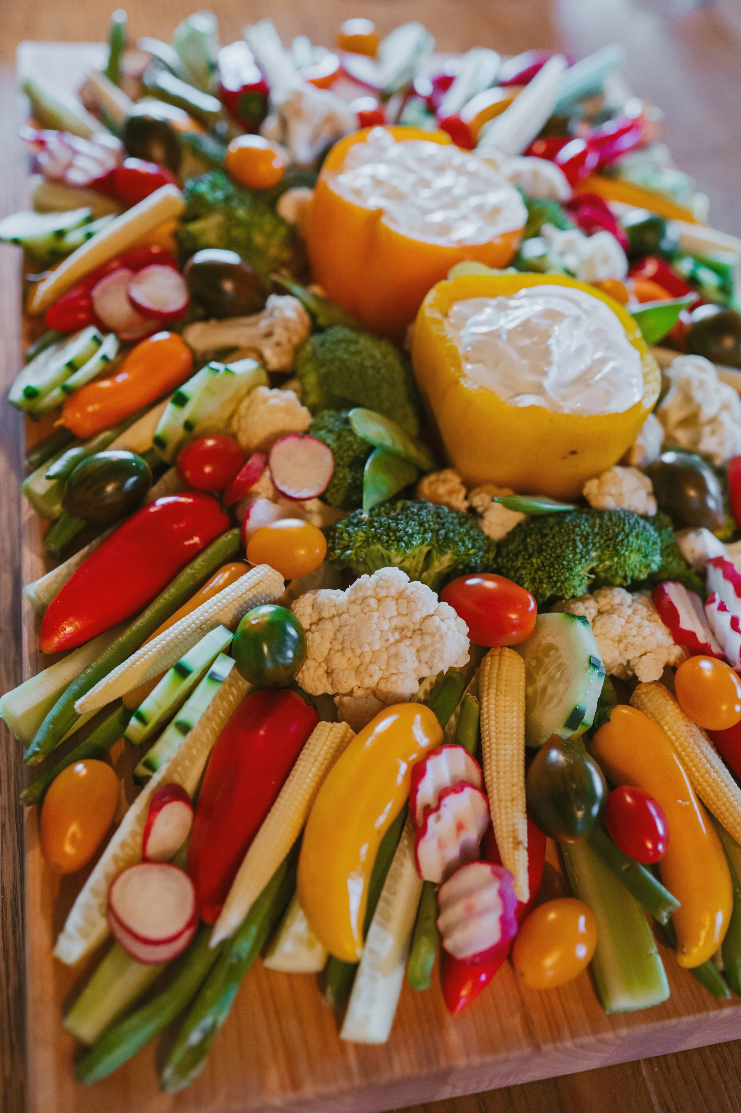 Colorful arrangement of raw vegetables including yellow and orange bell peppers, cherry tomatoes, cauliflower, broccoli, cucumbers, radishes, baby corn, and carrots on a wooden surface.