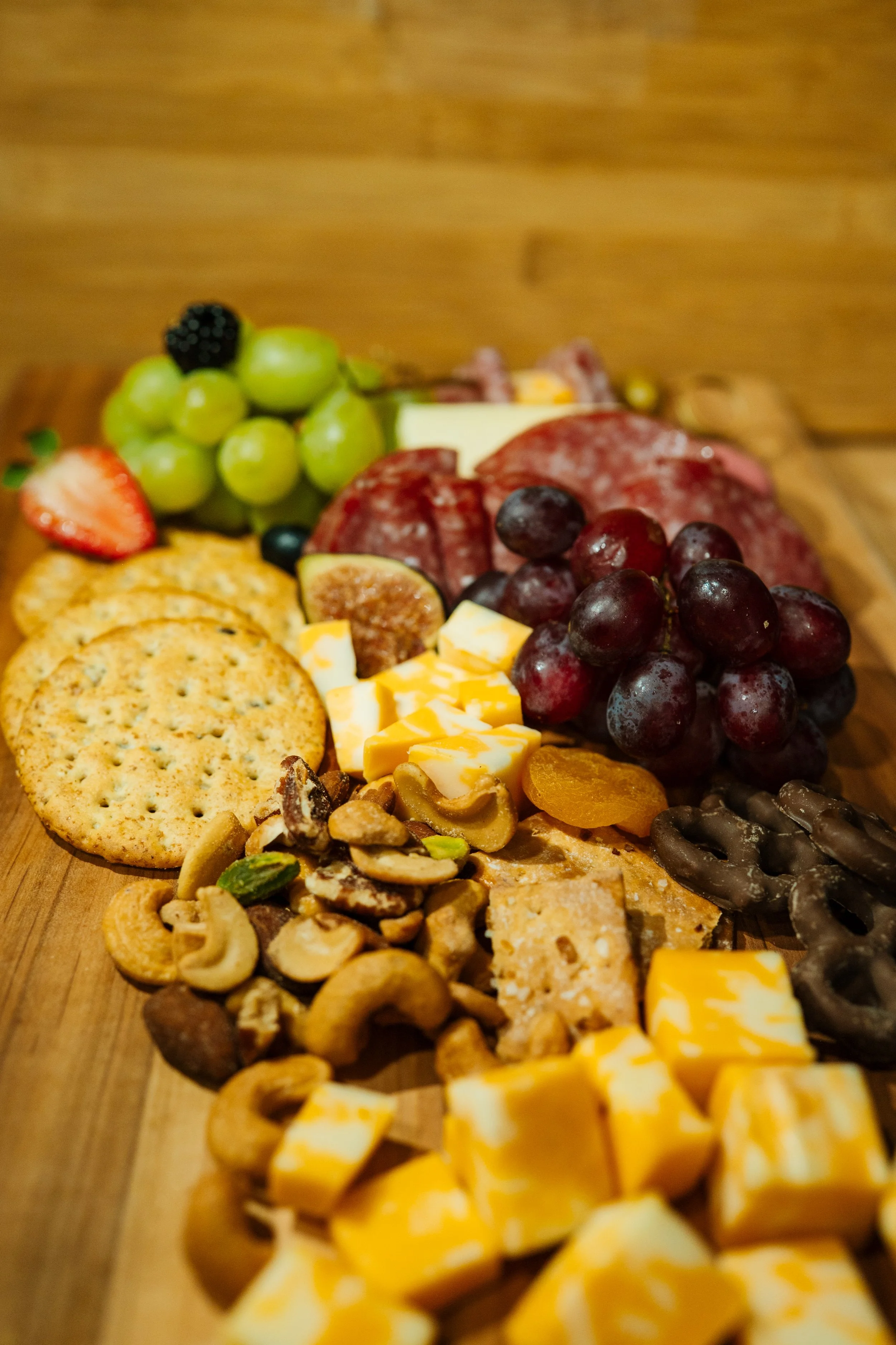 A variety of snacks including grapes, strawberries, blackberries, cheese cubes, crackers, nuts, dried fruit, and chocolate-covered pretzels arranged on a wooden surface.