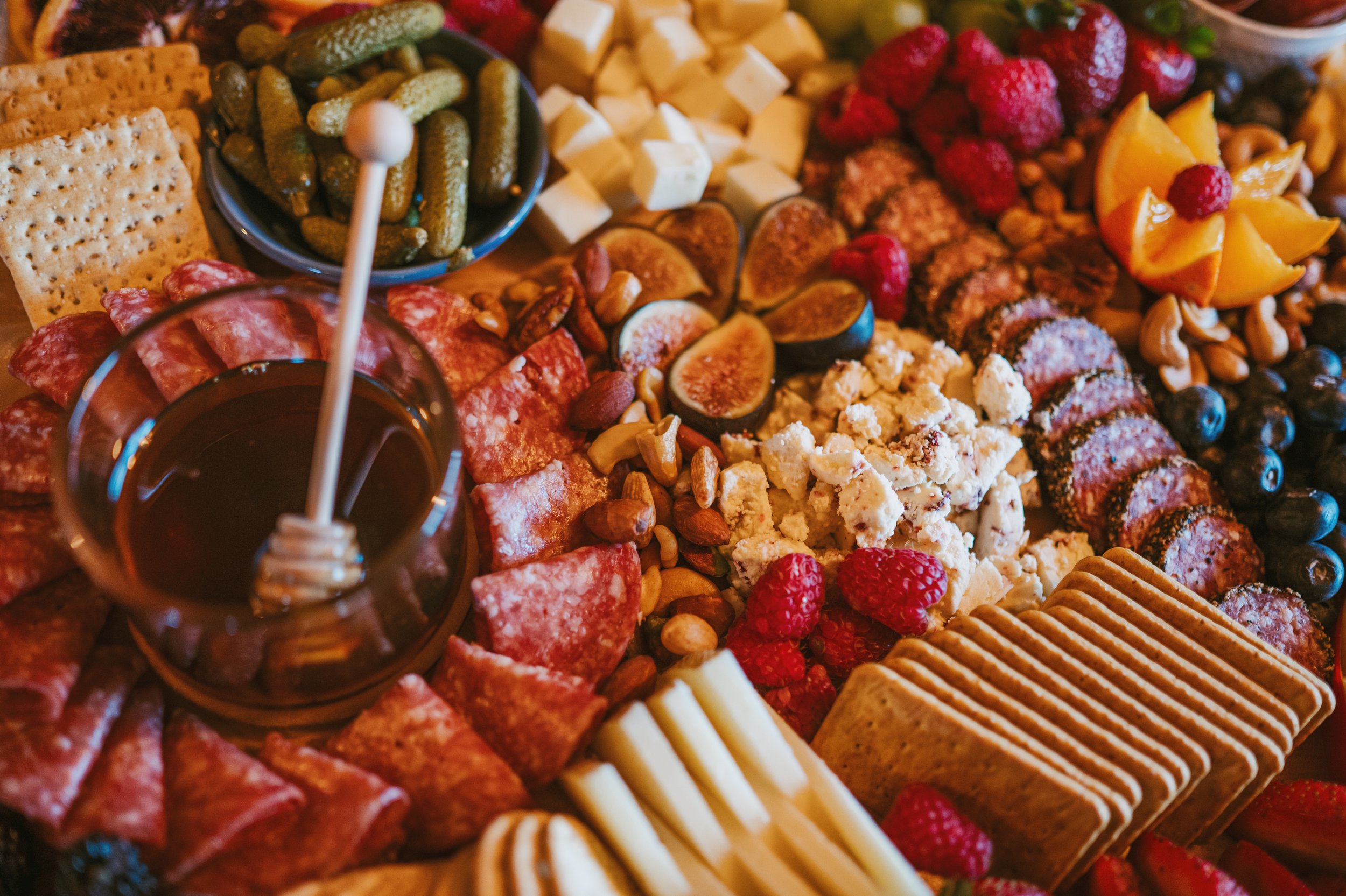 A variety of snacks including crackers, pickles, cheese cubes, strawberries, raspberries, blueberries, orange slices, nuts, and a small glass of red wine.