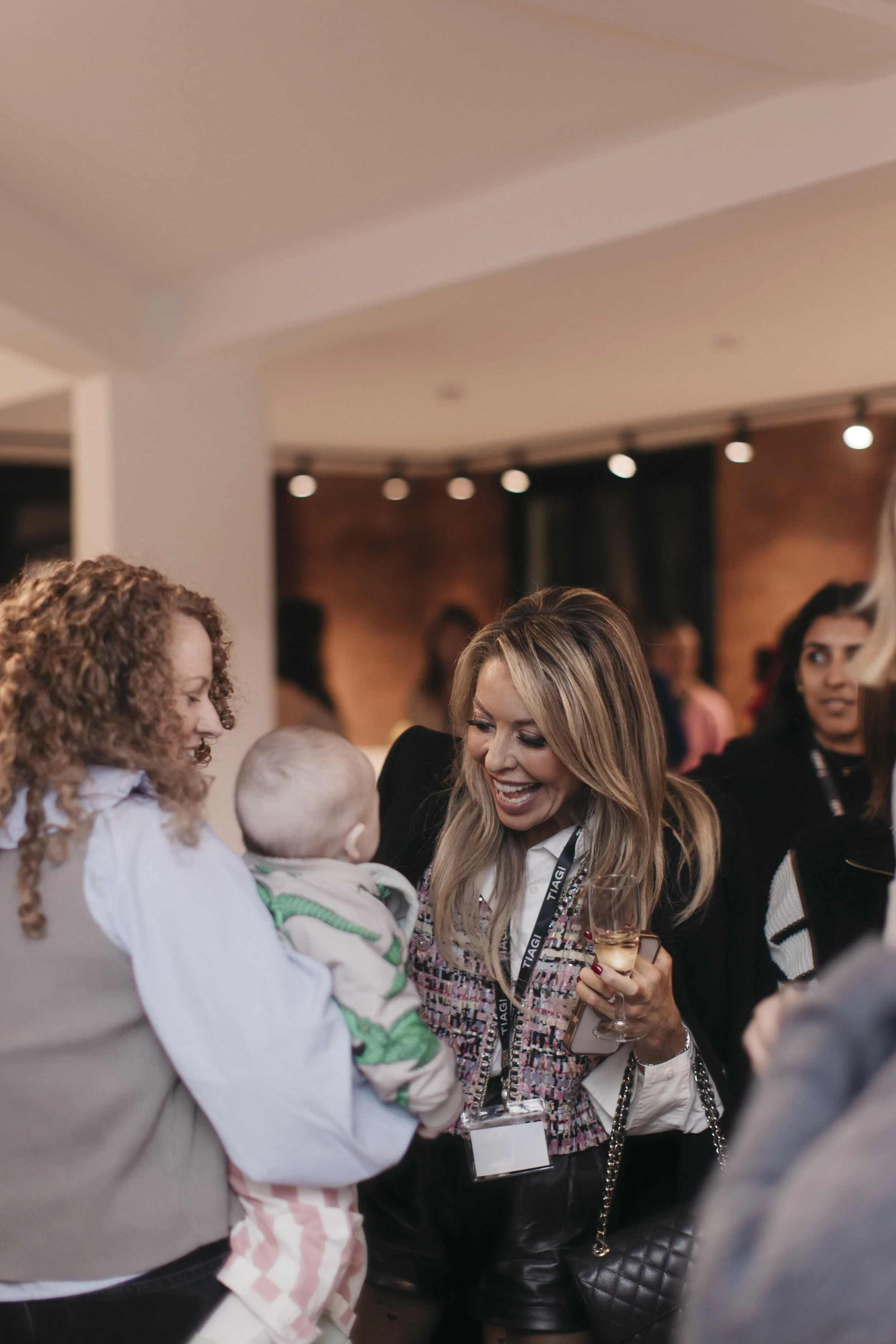 A woman with long blonde hair smiling and talking to a child held by another woman in a social gathering, holding a glass of wine.