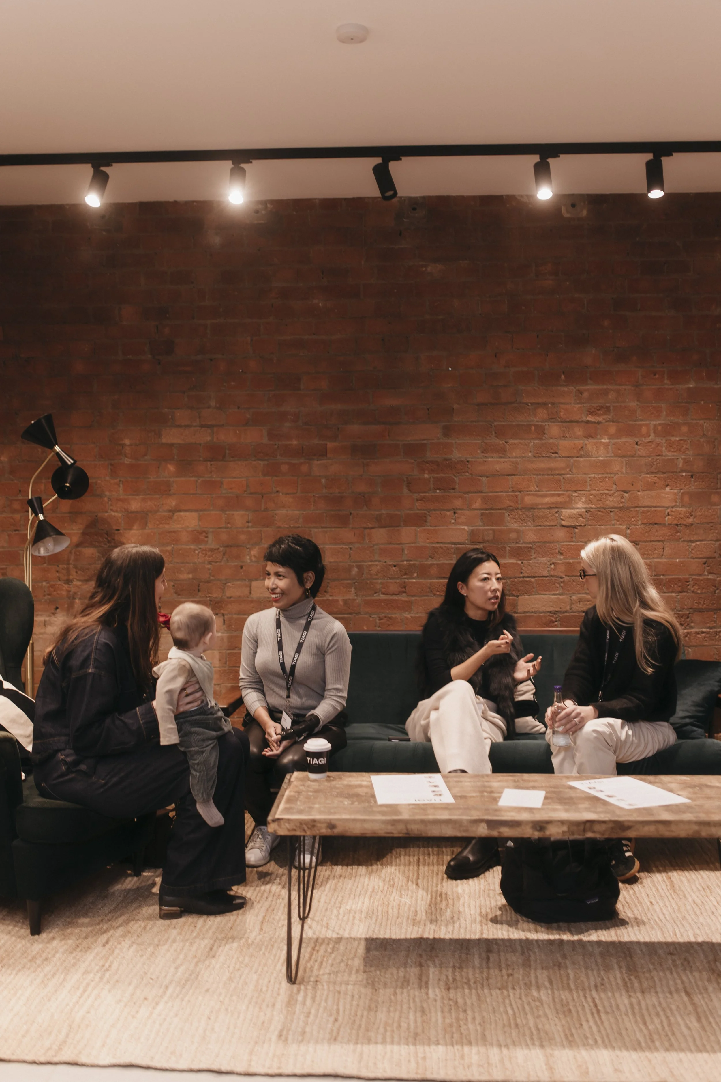 Four women, one holding a baby, sitting and talking on a dark green sofa against a brick wall in a casual indoor setting.