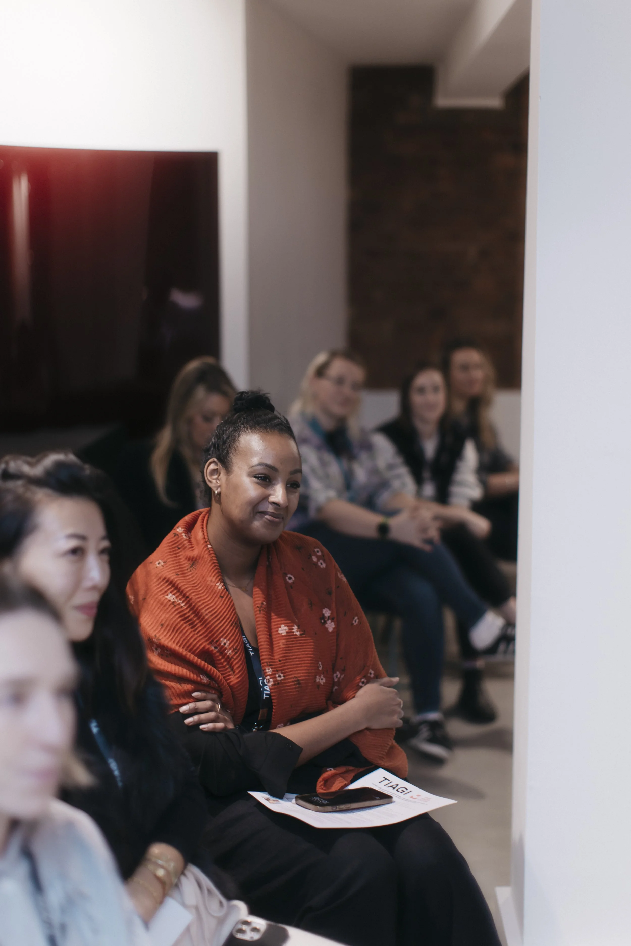 A diverse group of women attending a presentation or conference, sitting attentively in a room, with some holding papers and phones.