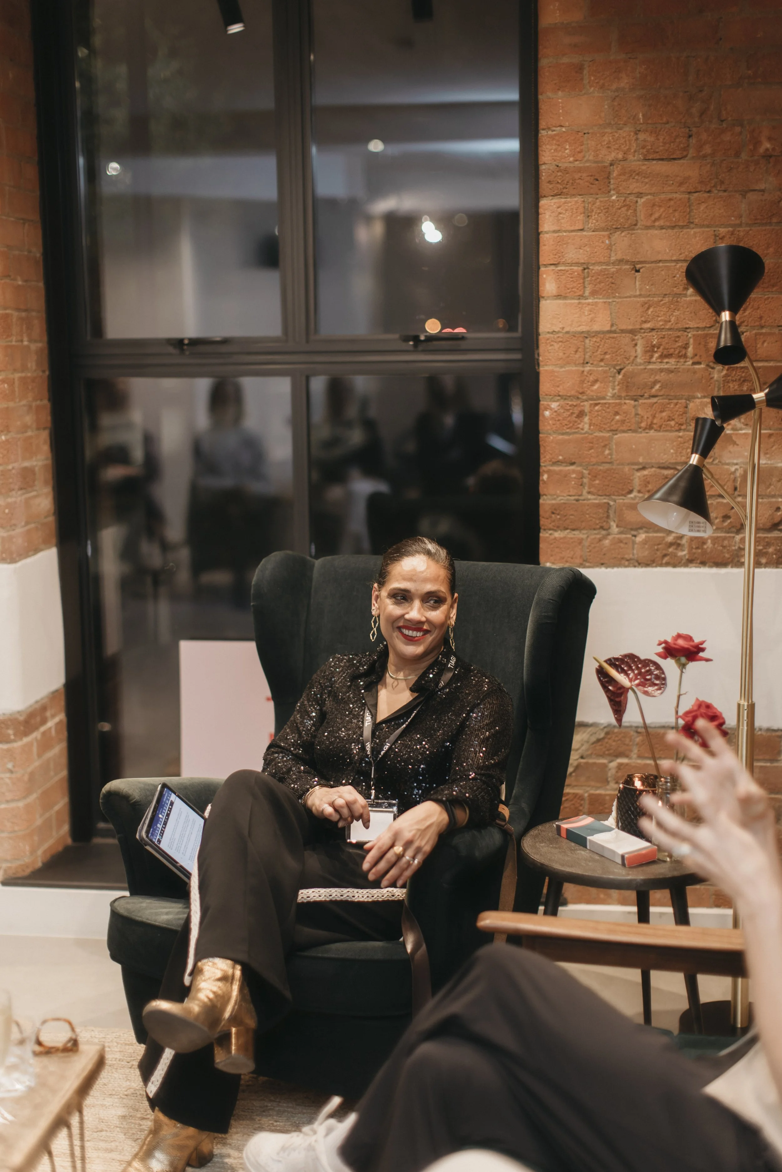 A woman sitting in a high-back black armchair, smiling, in a cozy indoor setting with a brick wall and large window behind her. She is wearing a black sequined top, black pants with gold details on the sides, and gold boots, with earrings and a watch. There is a side table with flowers and books beside her, and a person partially visible in the foreground, gesturing with their hand.