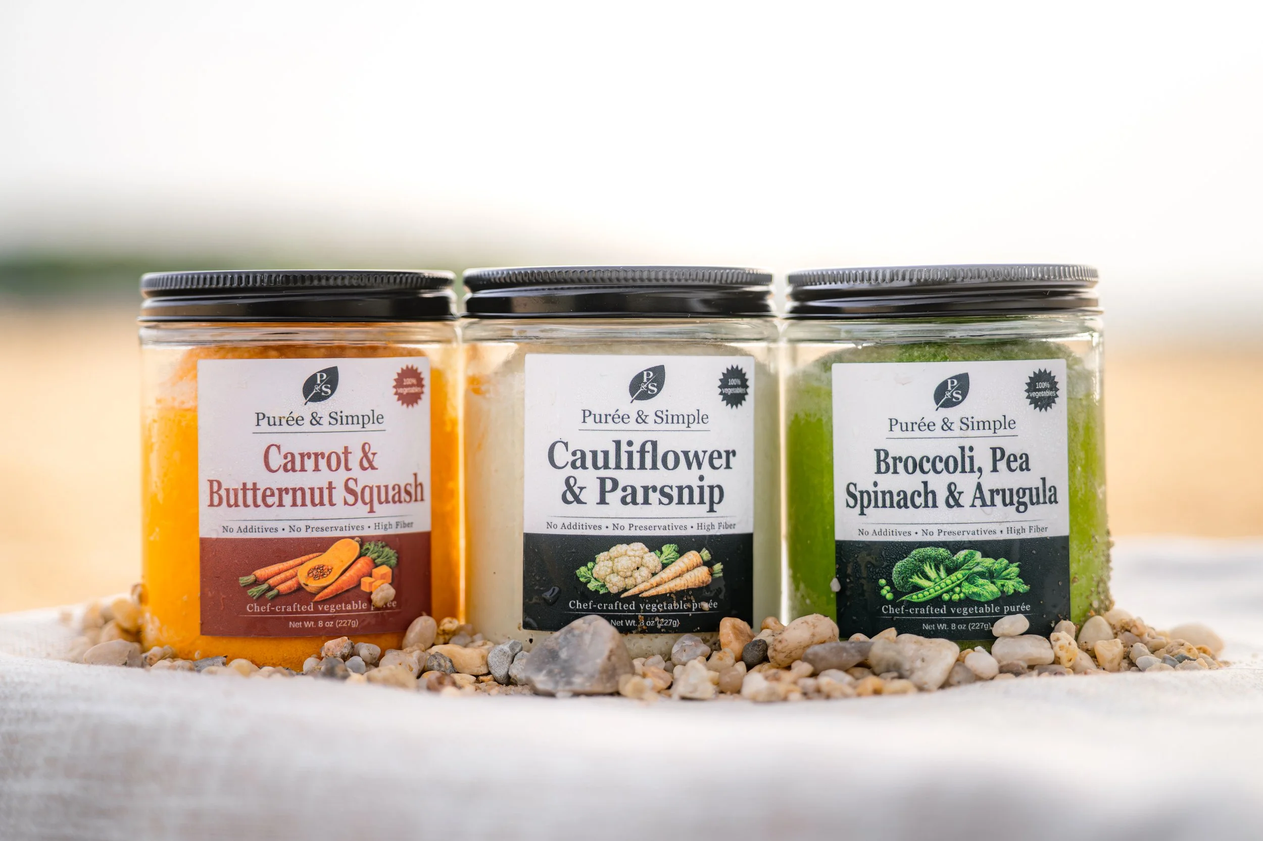 Three jars of vegetable purees labeled 'Carrot & Butternut Squash,' 'Cauliflower & Parsnip,' and 'Broccoli, Pea, Spinach & Arugula,' placed on a surface with small pebbles.