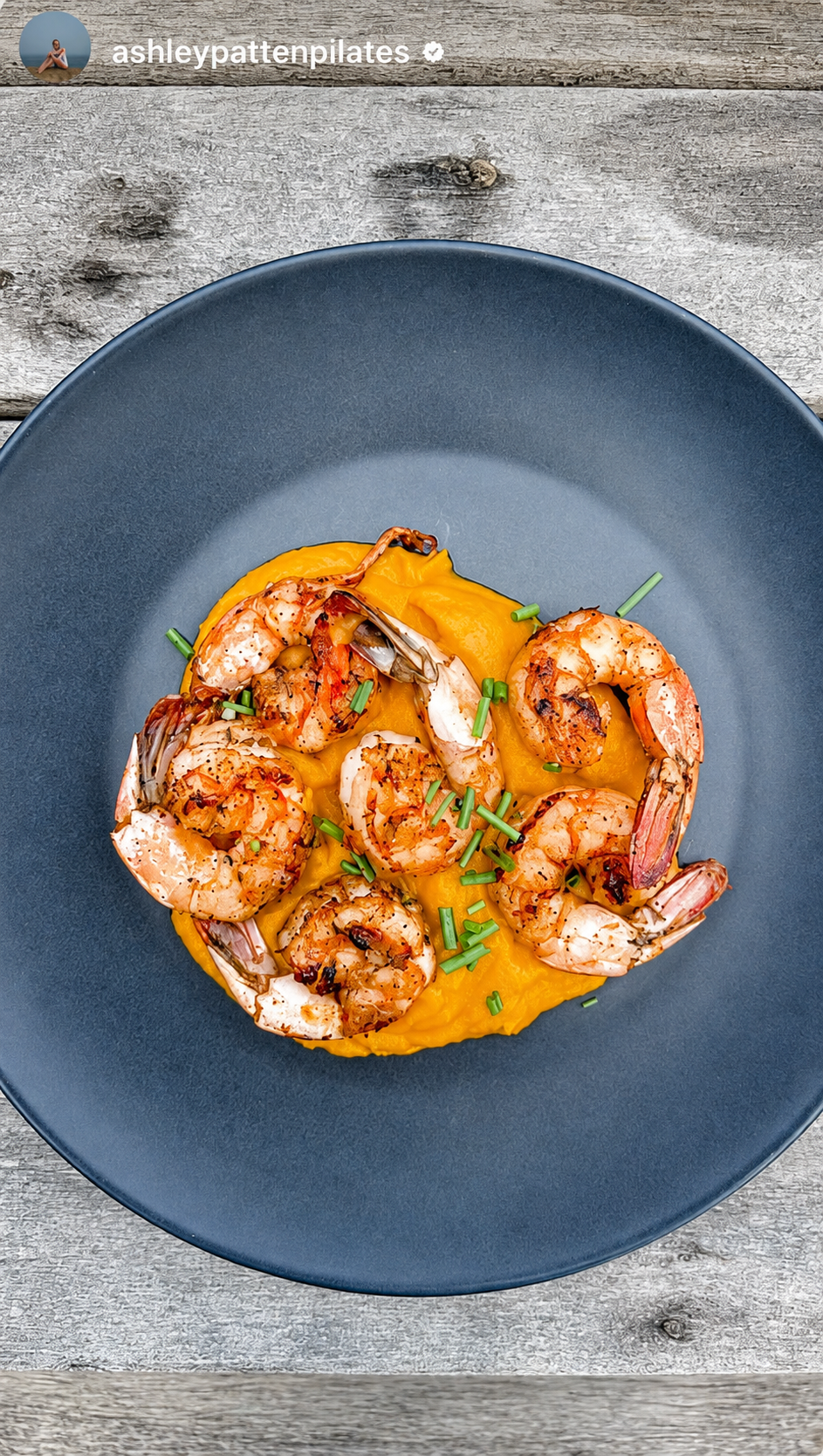 A black plate with grilled shrimp served over bright yellow mashed sweet potatoes, garnished with chopped chives, placed on a rustic wooden table.