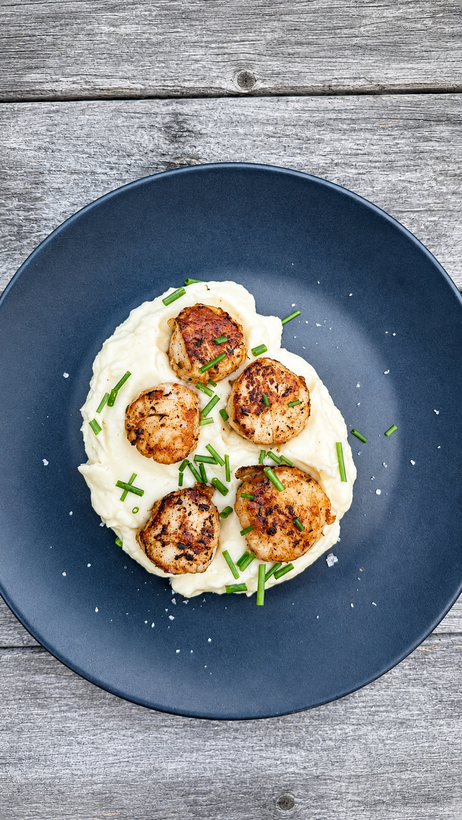Seared scallops served on a bed of creamy mashed potatoes, garnished with chopped chives on a dark plate.
