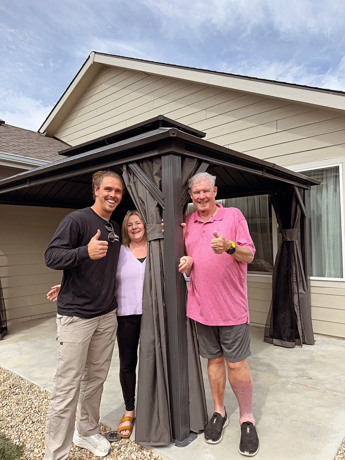 Three people standing outside of a house, smiling and giving thumbs up, next to a new outdoor gazebo with curtains.