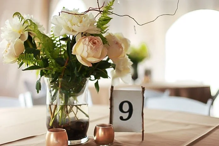 A floral centerpiece with white and pale pink roses and green foliage in a glass vase, accompanied by a table number sign with the number 9 and two small metallic candle holders on a beige tablecloth at a banquet or wedding reception.