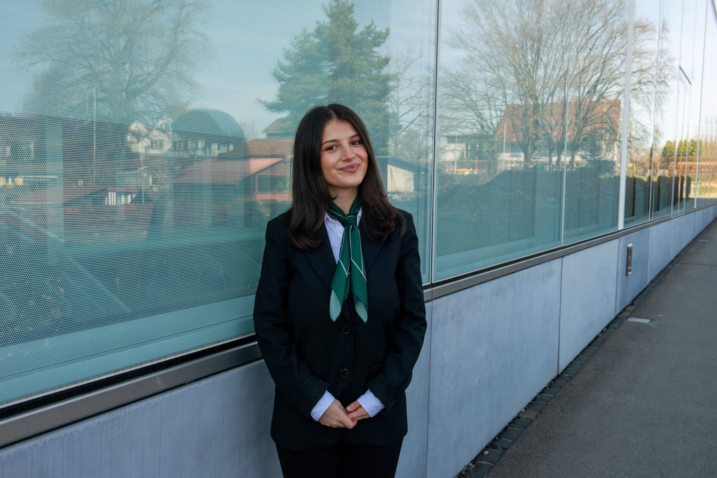 A young woman in a dark suit with a green and white scarf standing in front of a glass building with trees reflecting on the glass.