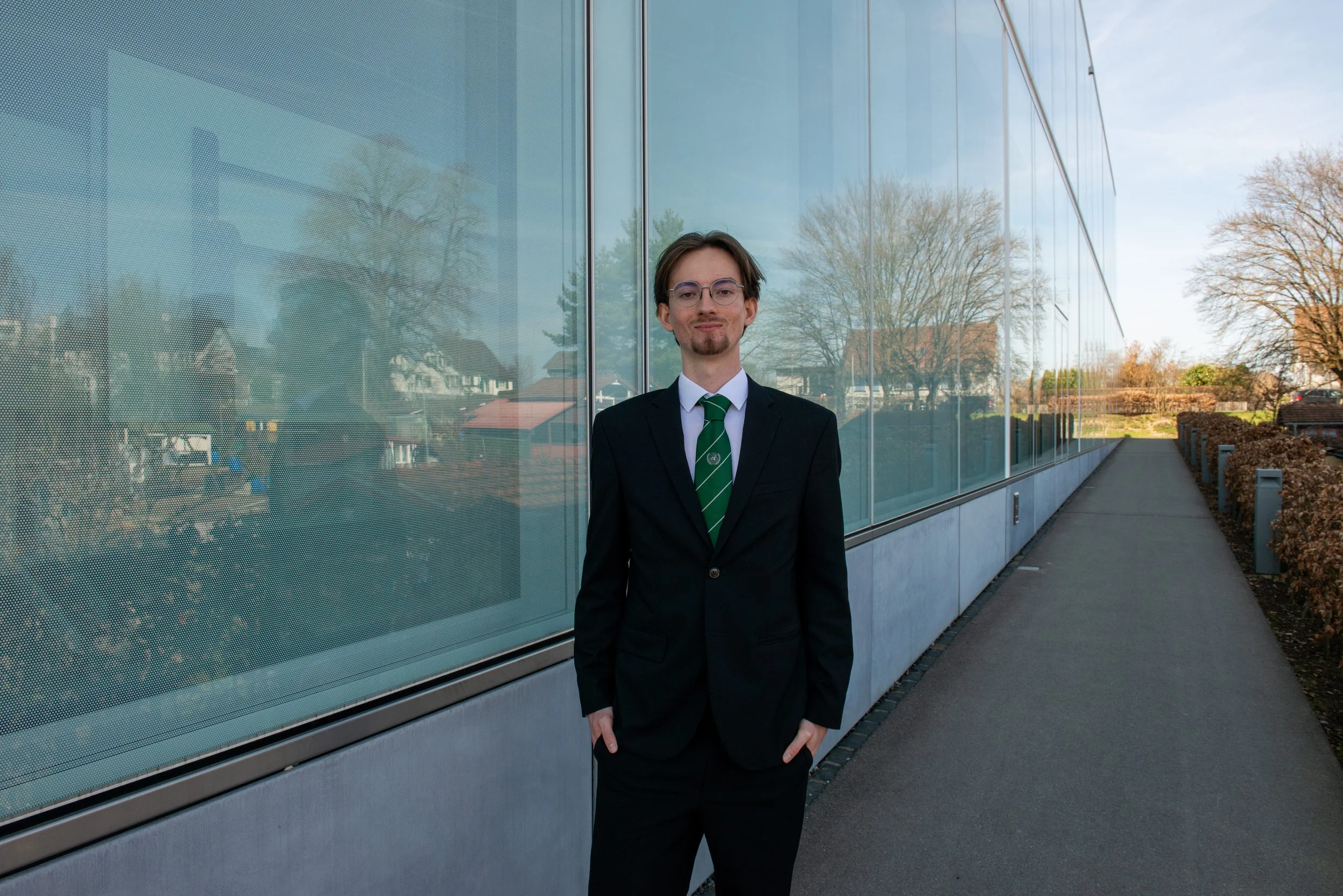 A young man with brown hair, glasses, and a goatee, wearing a black suit, white shirt, and green striped tie, standing outside next to a modern glass building.