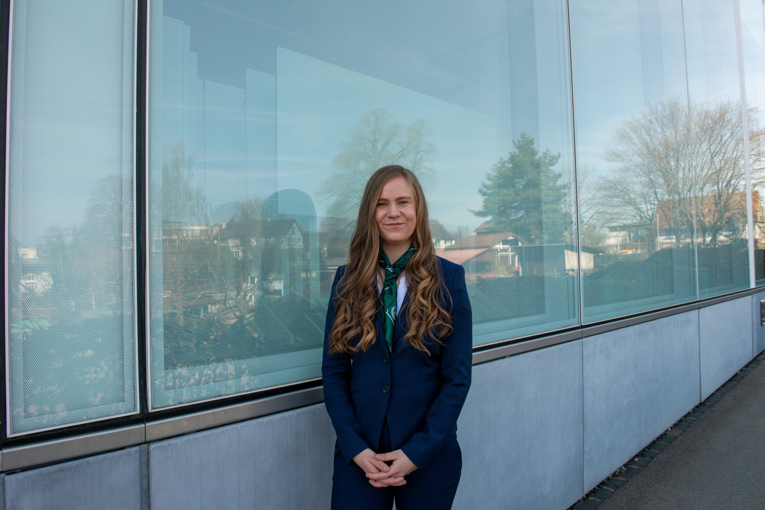 A young woman in a navy blazer, white shirt, and a green scarf tied around her neck, standing outside in front of a building with large glass windows, smiling at the camera.