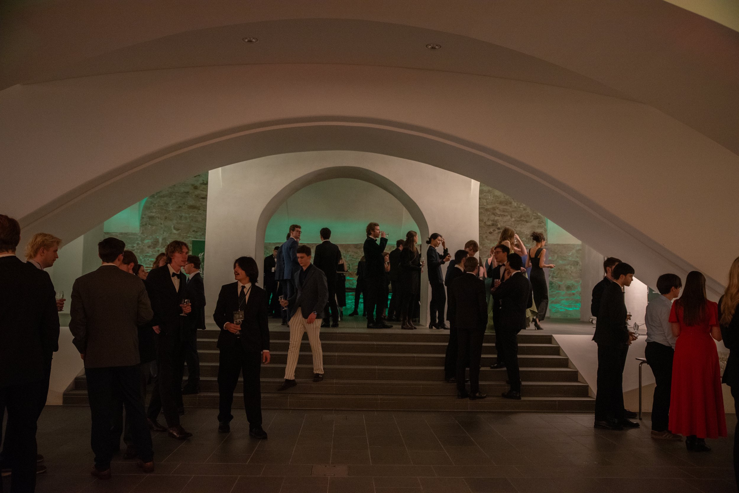 People in formal attire at a social event in an elegant venue with arched architecture and steps leading to a raised platform.