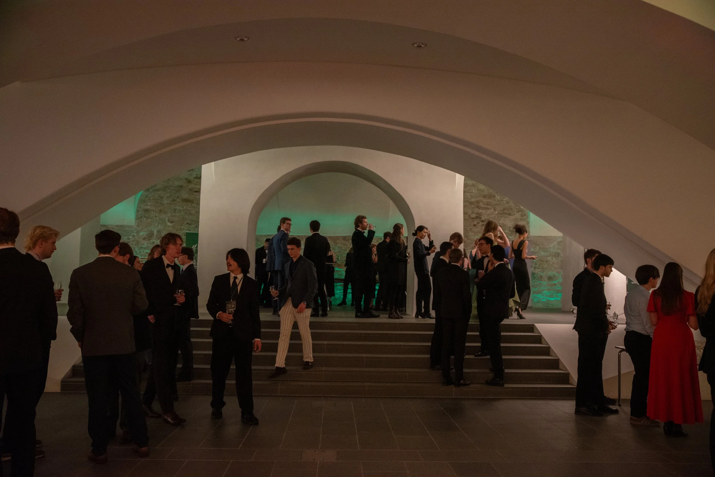 People socializing at a formal event or reception in an elegant, modern venue with arched architecture and green accent lighting.