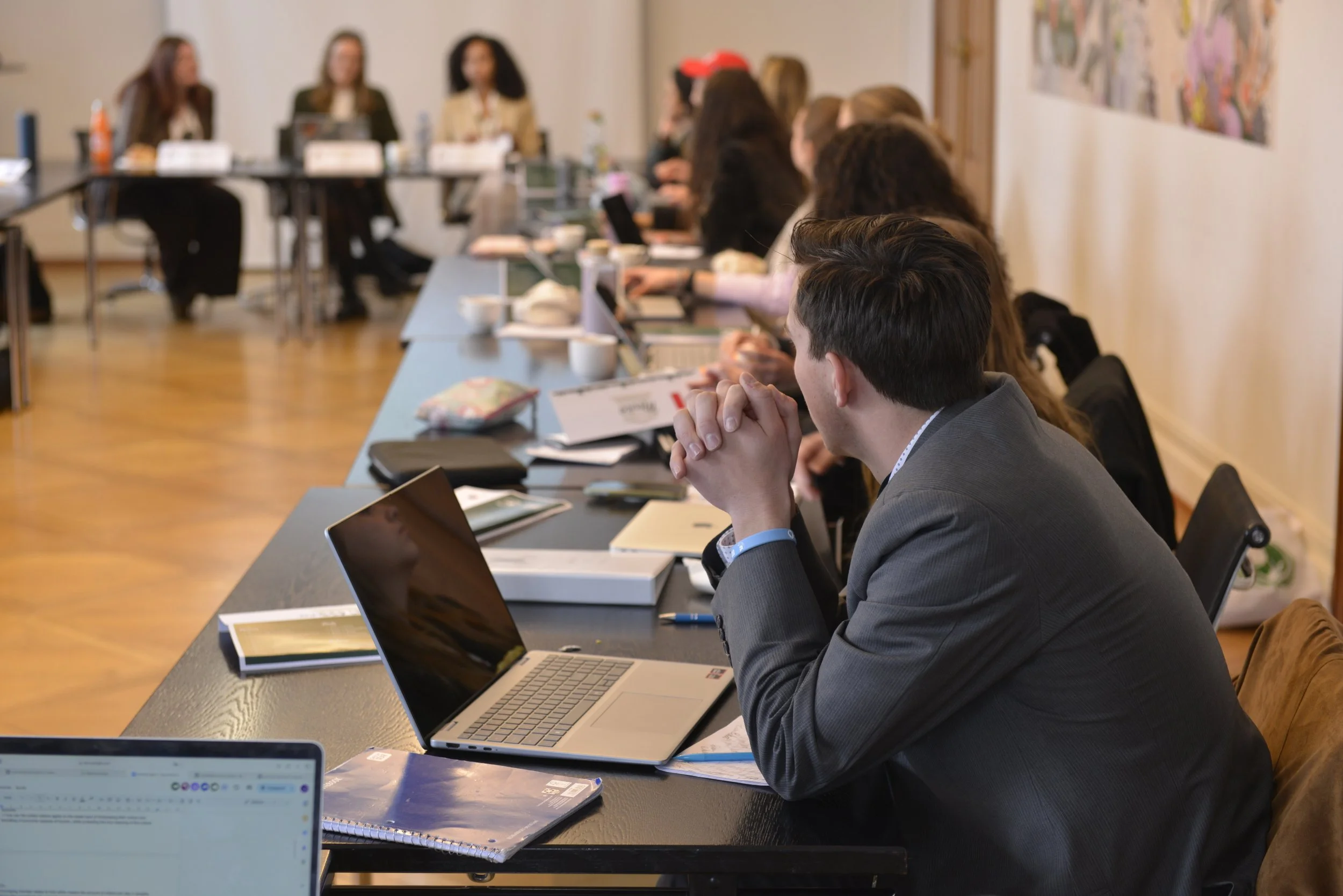 A man in a gray suit sitting at a conference table with a silver laptop, with several other people in the background attending a meeting in a room with framed artwork on the wall.
