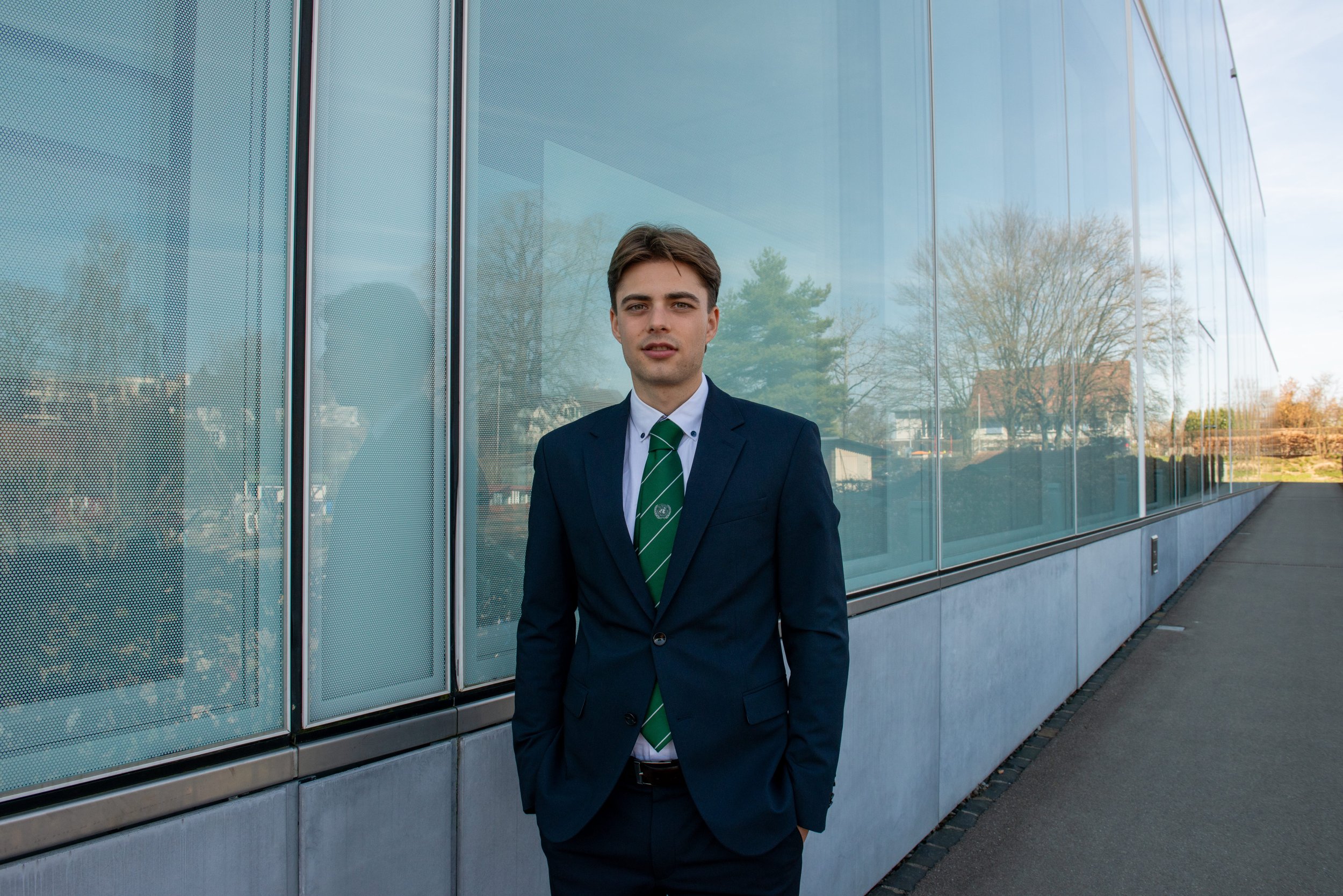 Young man in a navy suit and green striped tie standing outside next to a modern glass building during daytime.