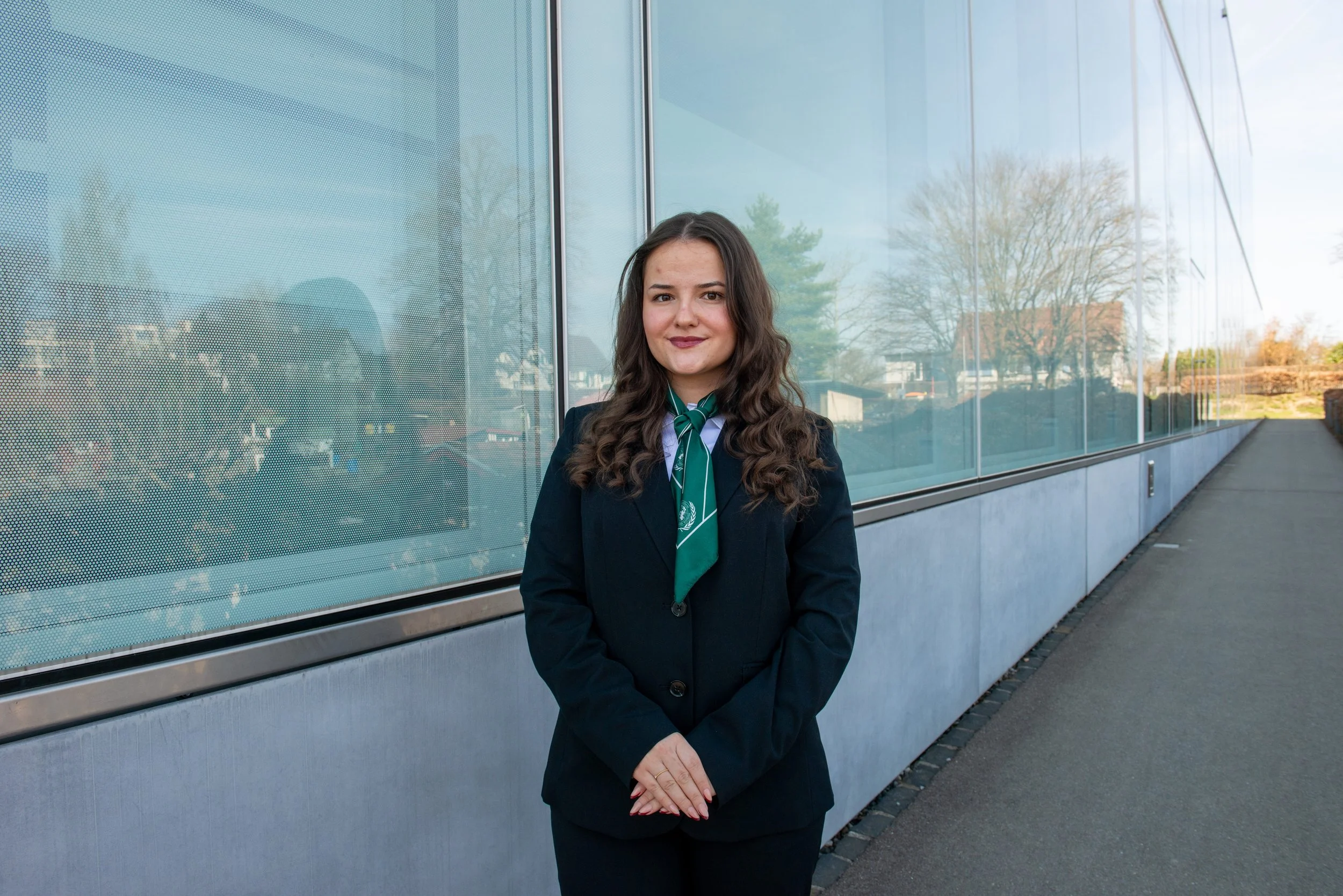 Woman in formal business attire with a green scarf standing outside next to a modern glass building.