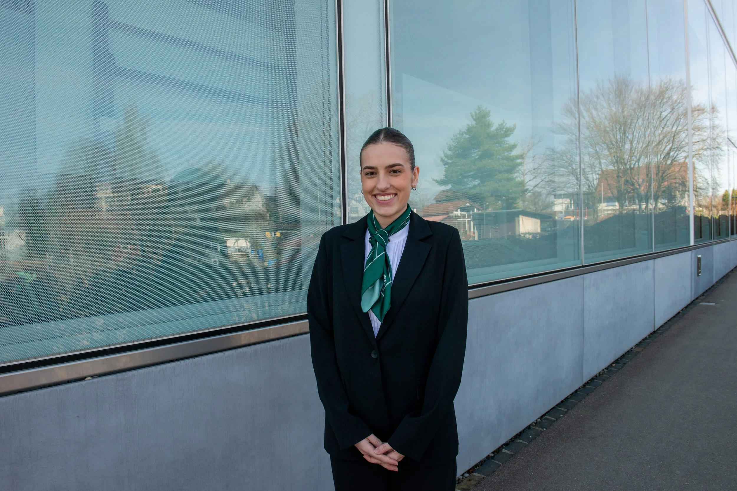 A young woman in formal business attire with a green scarf, standing outside in front of a modern glass building, smiling at the camera.