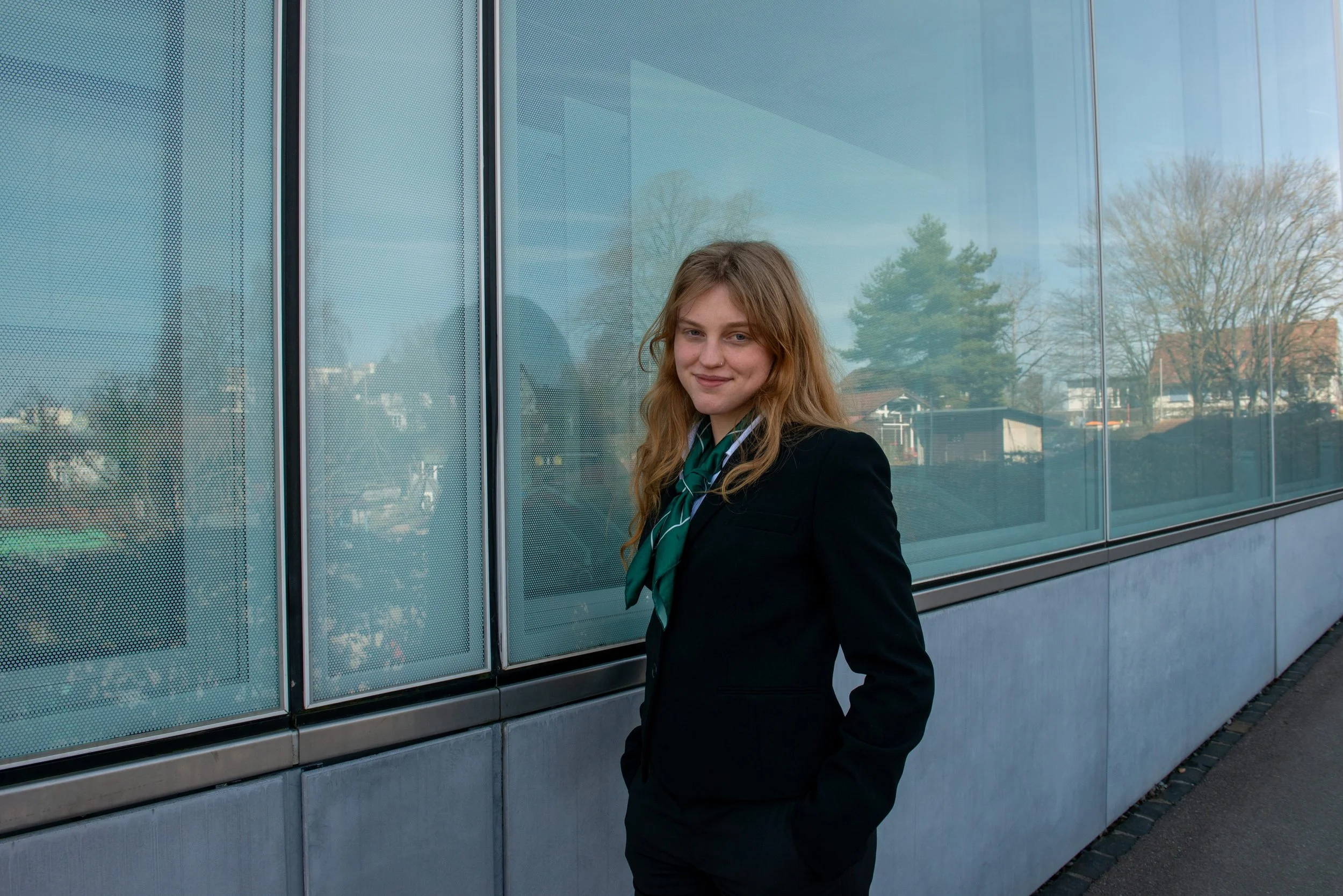 A young woman with long wavy red hair, wearing a black blazer, white shirt, and a green scarf, standing outdoors in front of a glass building with reflections of trees and houses.