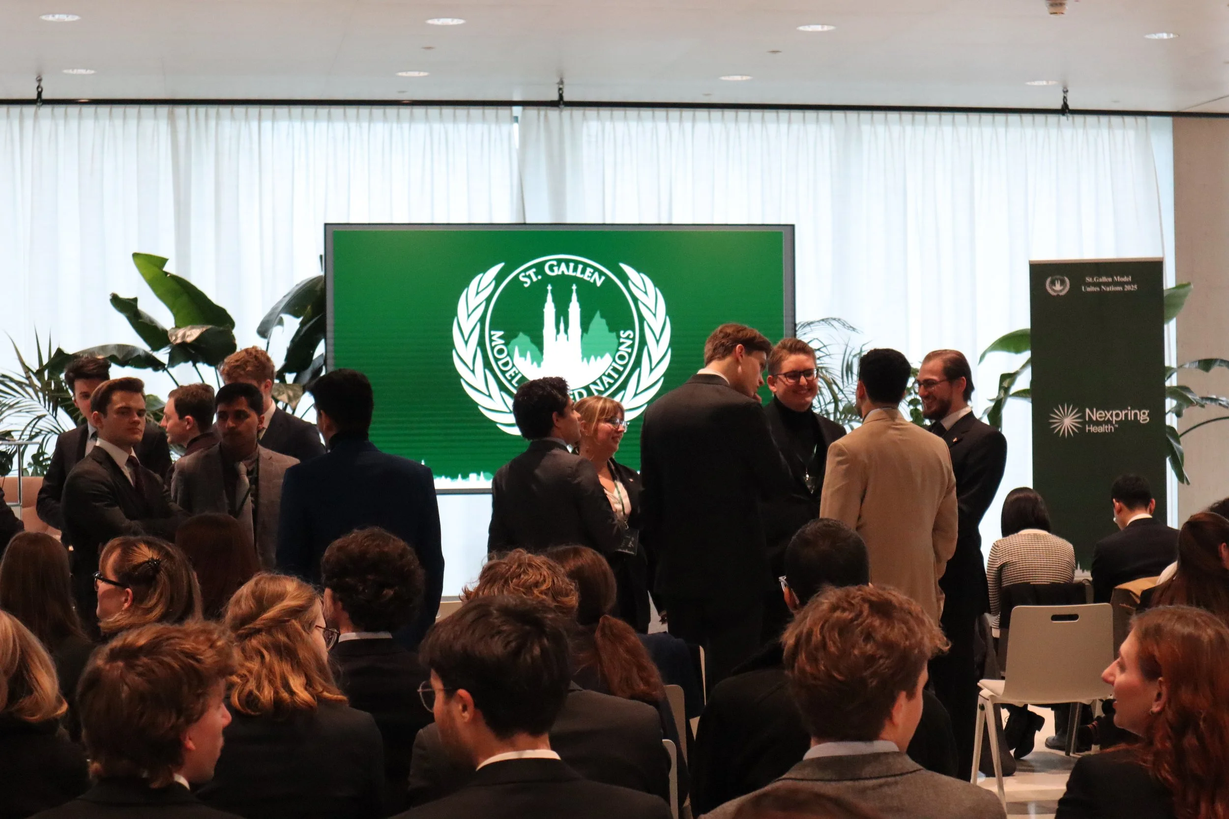 Group of professionals in formal attire gathered at a conference or networking event, with a large display showing the logo of St. Gallen Model United Nations in the background.
