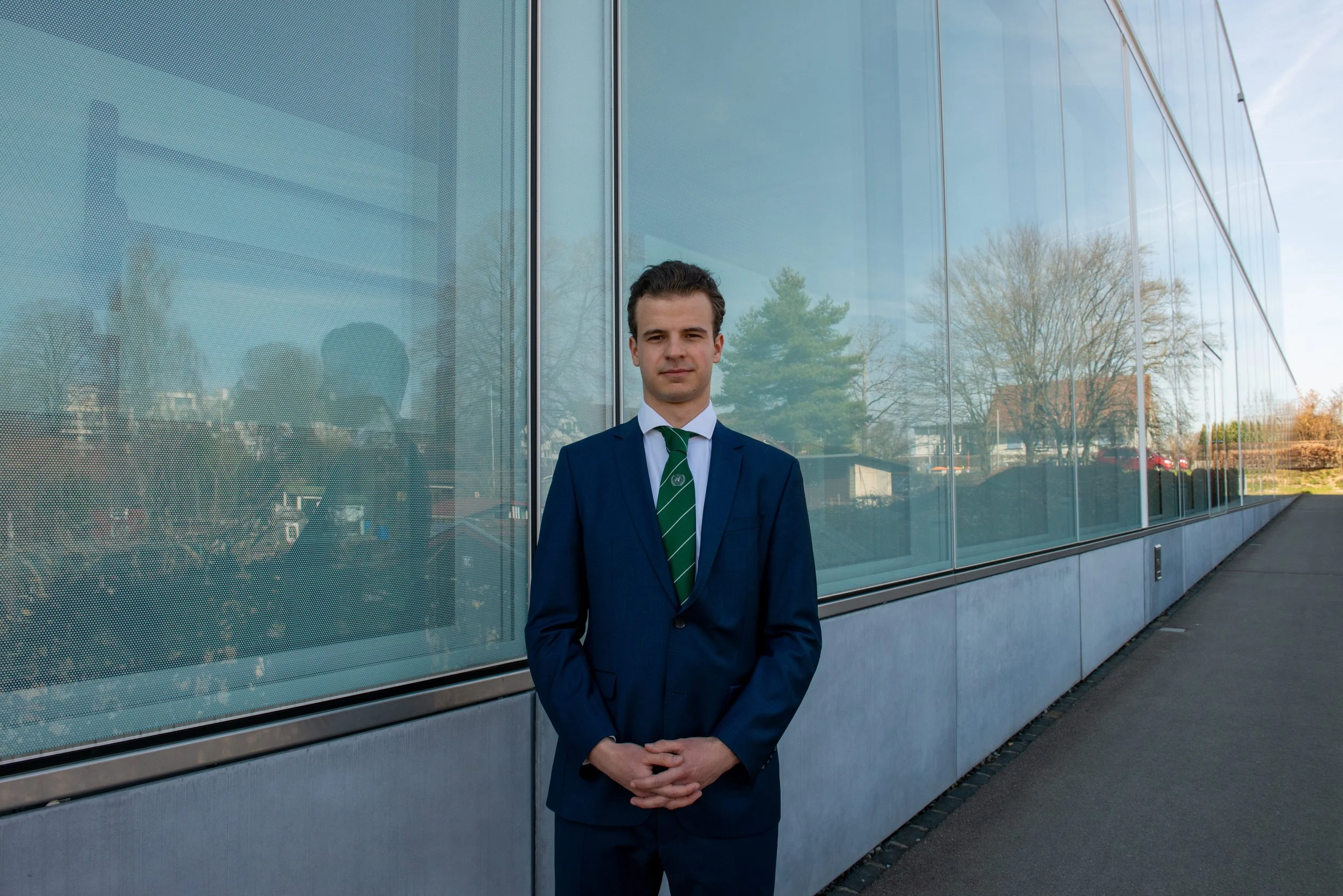 A man in a navy blue suit, white shirt, and green striped tie, standing outside in front of a reflective glass building, with trees and a partly cloudy sky in the background.
