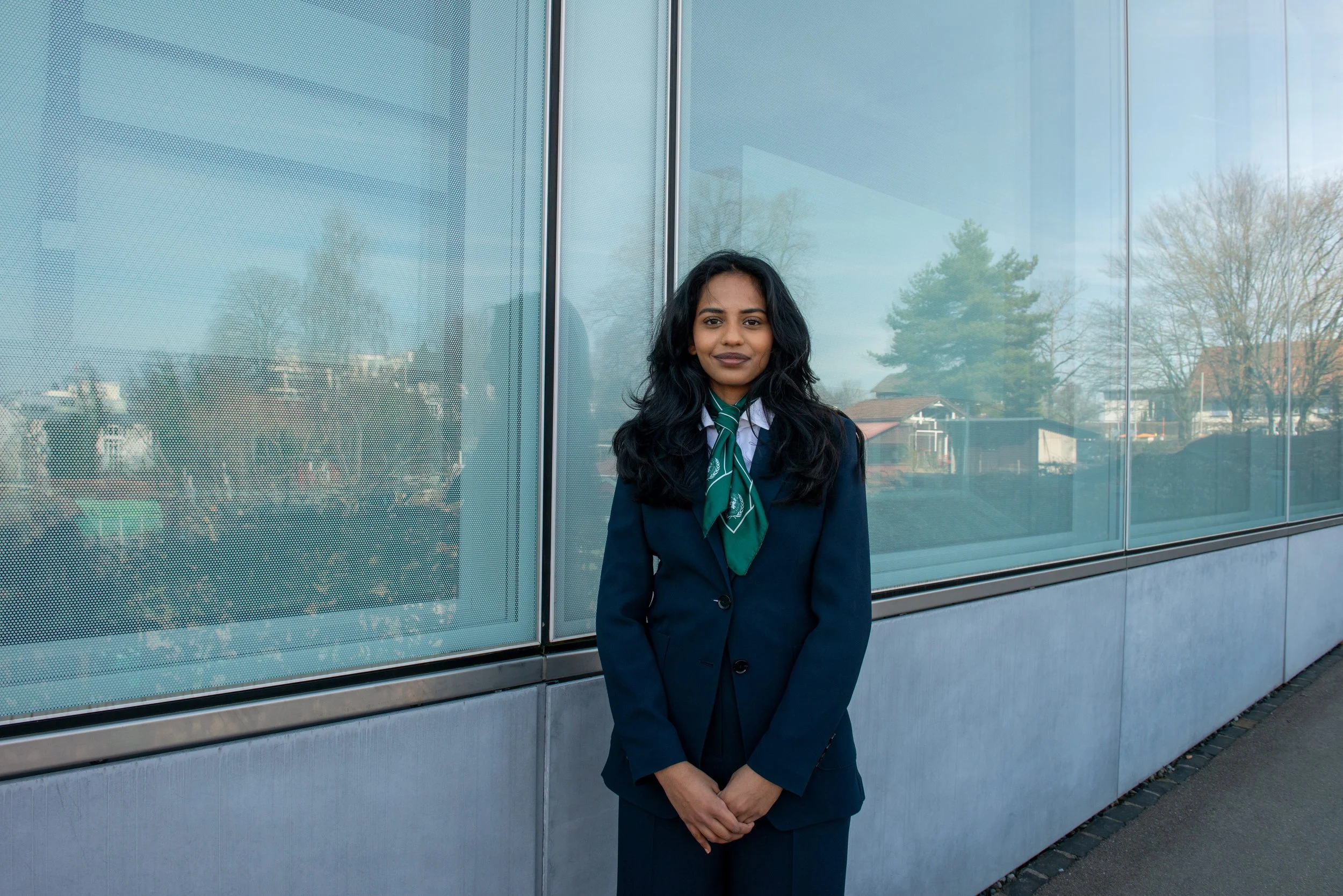 A woman in a navy blazer and green necktie standing outdoors in front of a glass building with reflections of trees and houses.
