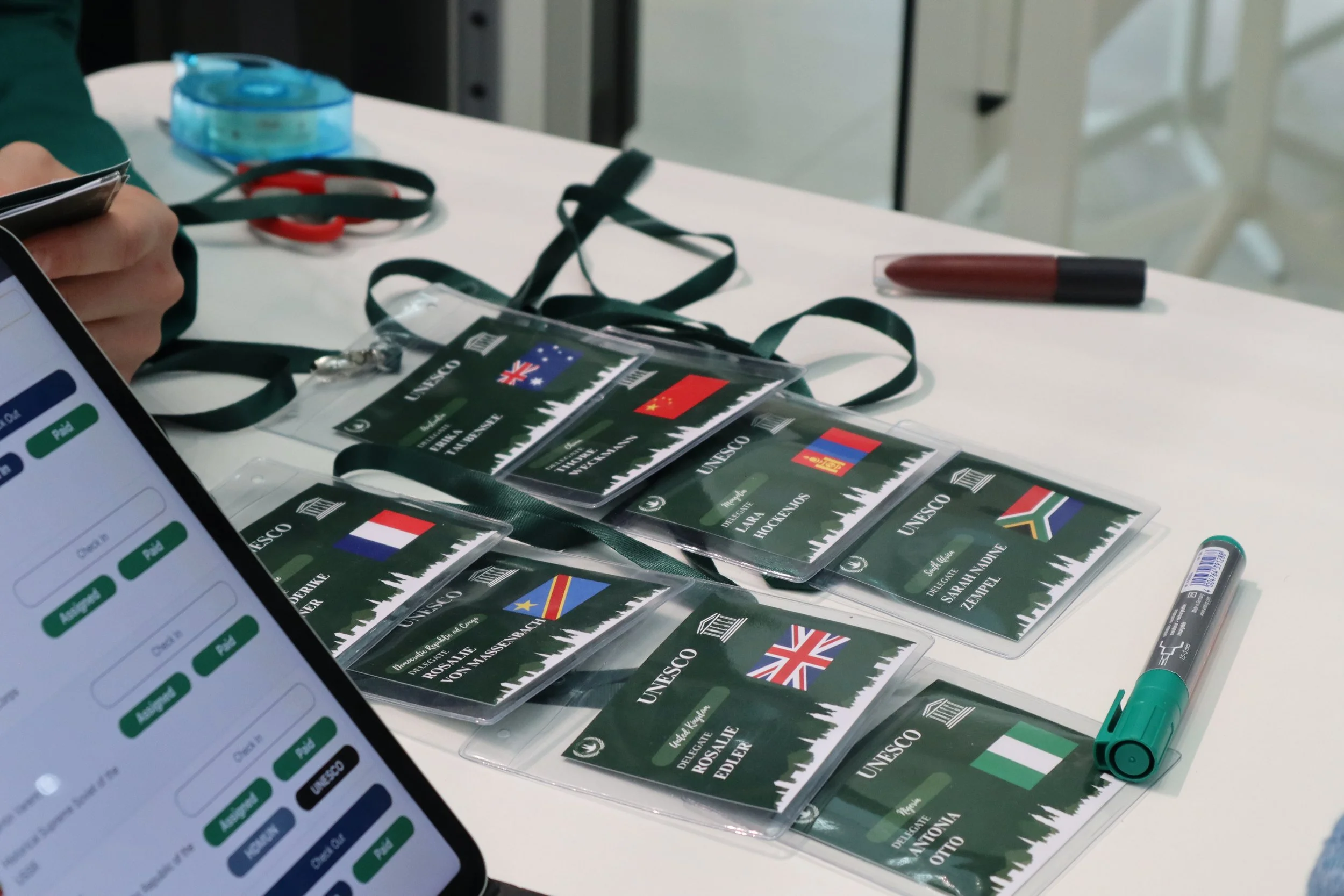 Name badges with the UNESCO logo and flags of Australia, China, Netherlands, South Africa, and Italy on a white table, along with a green marker, a black marker, a smartphone, and a marker cap.