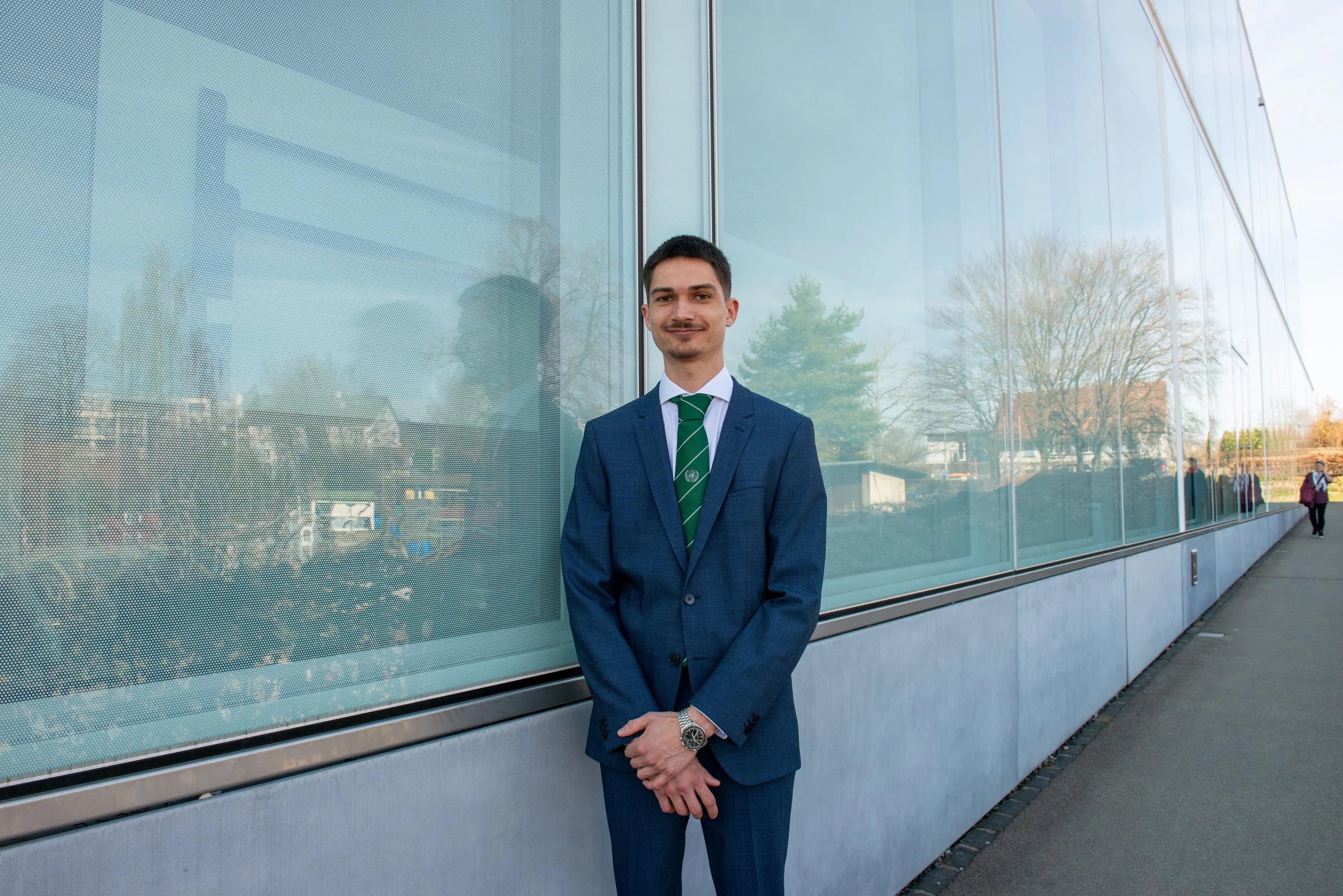 A man in a blue suit and green striped tie standing outdoors near a modern building with reflective glass panels.