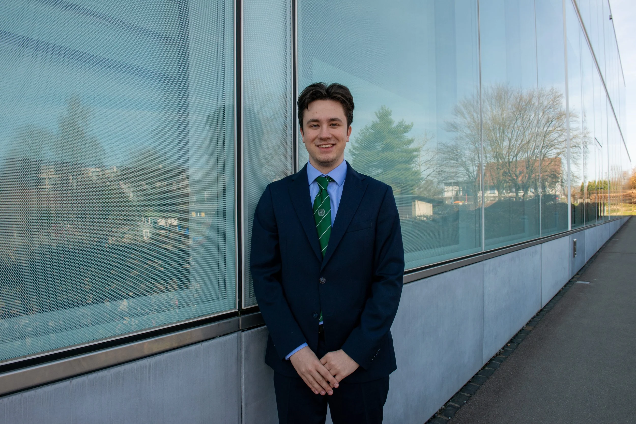 Young man in a navy suit and green striped tie standing outside near a large glass building, smiling and looking at the camera.