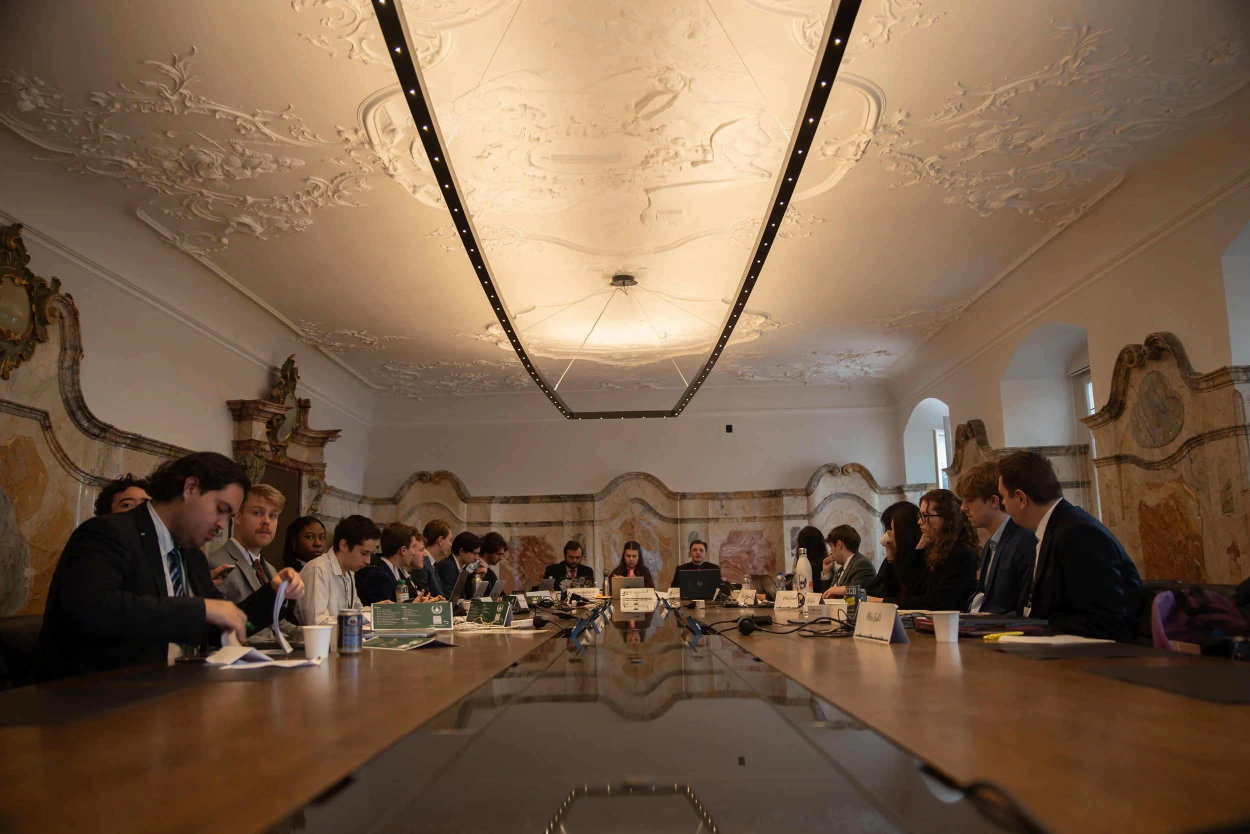 A formal meeting in a grand, ornate room with high ceilings and decorative molding, featuring around 20 people seated around a long wooden table, some with laptops and documents, engaging in discussion.