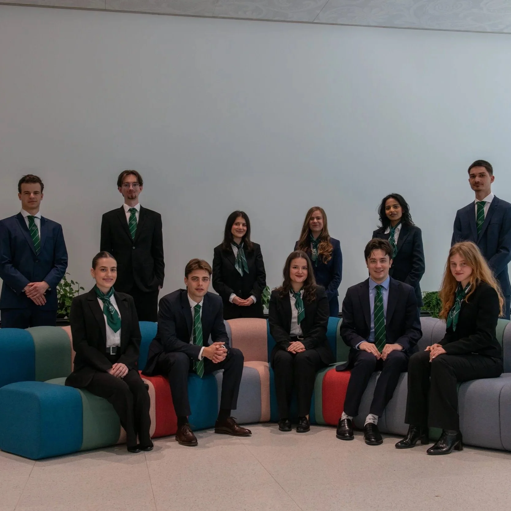 Group of ten young professionals in formal business attire, sitting and standing in a modern office space with a plain white wall and colorful seating.