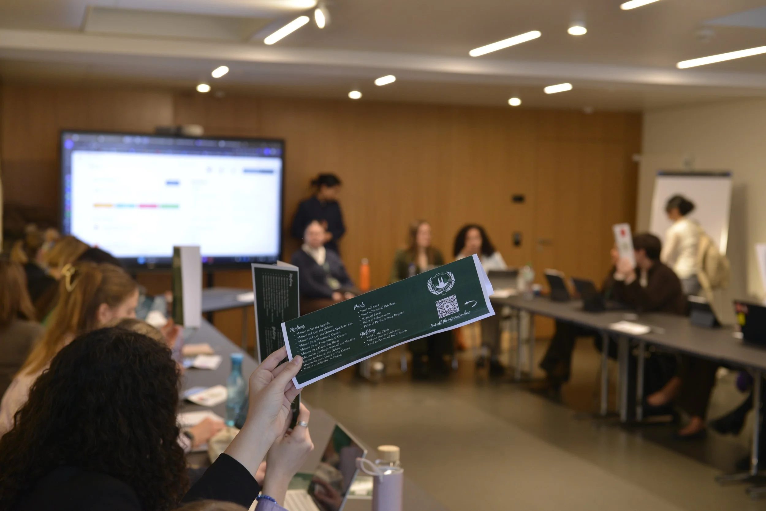 Person holding a brochure in a conference room with people seated at a long table and a large screen at the front.