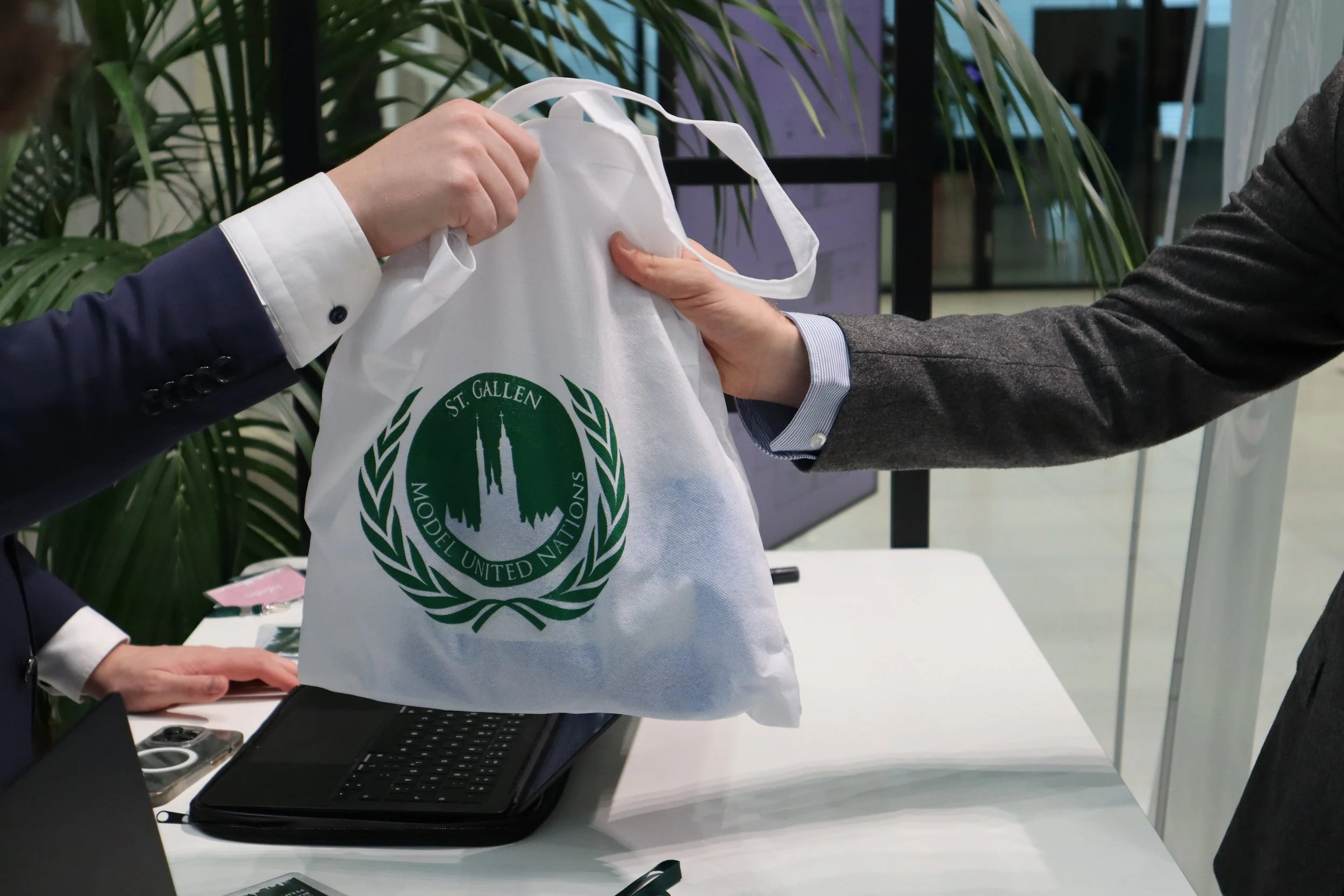 Two men in business suits exchanging a white bag with the logo of St. Gallen Model United Nations, at a desk with a laptop and papers, with green plants in the background.