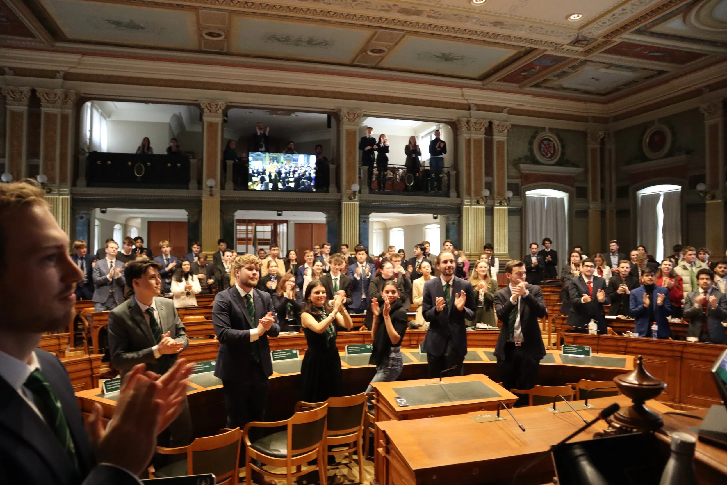 People in formal attire standing and clapping in a large ornate hall with high ceiling and decorative details, some on a balcony, during an event or ceremony.