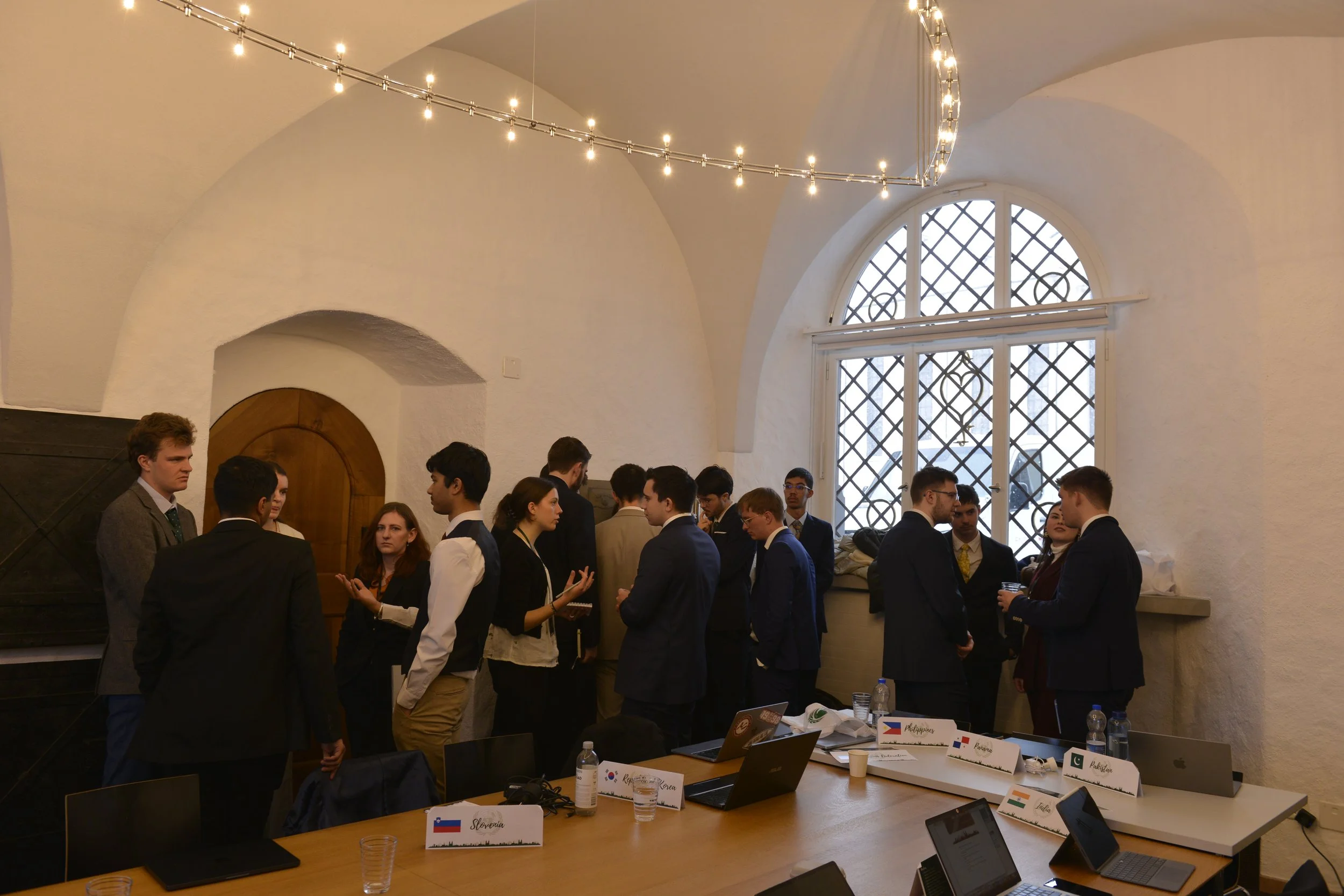 Group of young professionals engaging in discussions in a conference room with a large window and minimal decor.