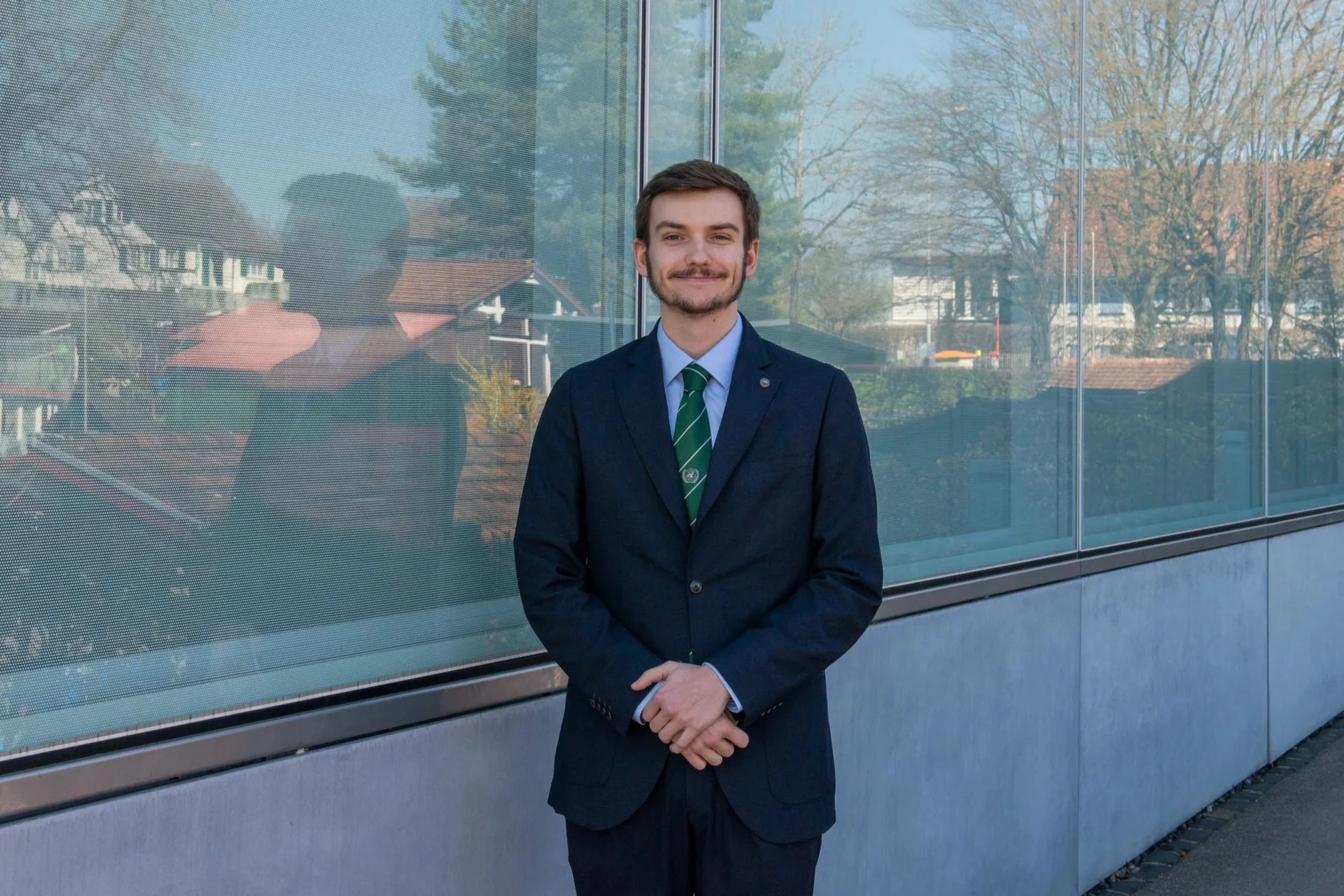 Young man in a navy suit and green striped tie stands in front of a modern glass building, with reflections of trees and houses in the background.