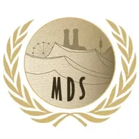A circular emblem with a beige background, featuring a bridge, mountains, and a cable car, framed by a laurel wreath, with the initials 'MDS' at the bottom.
