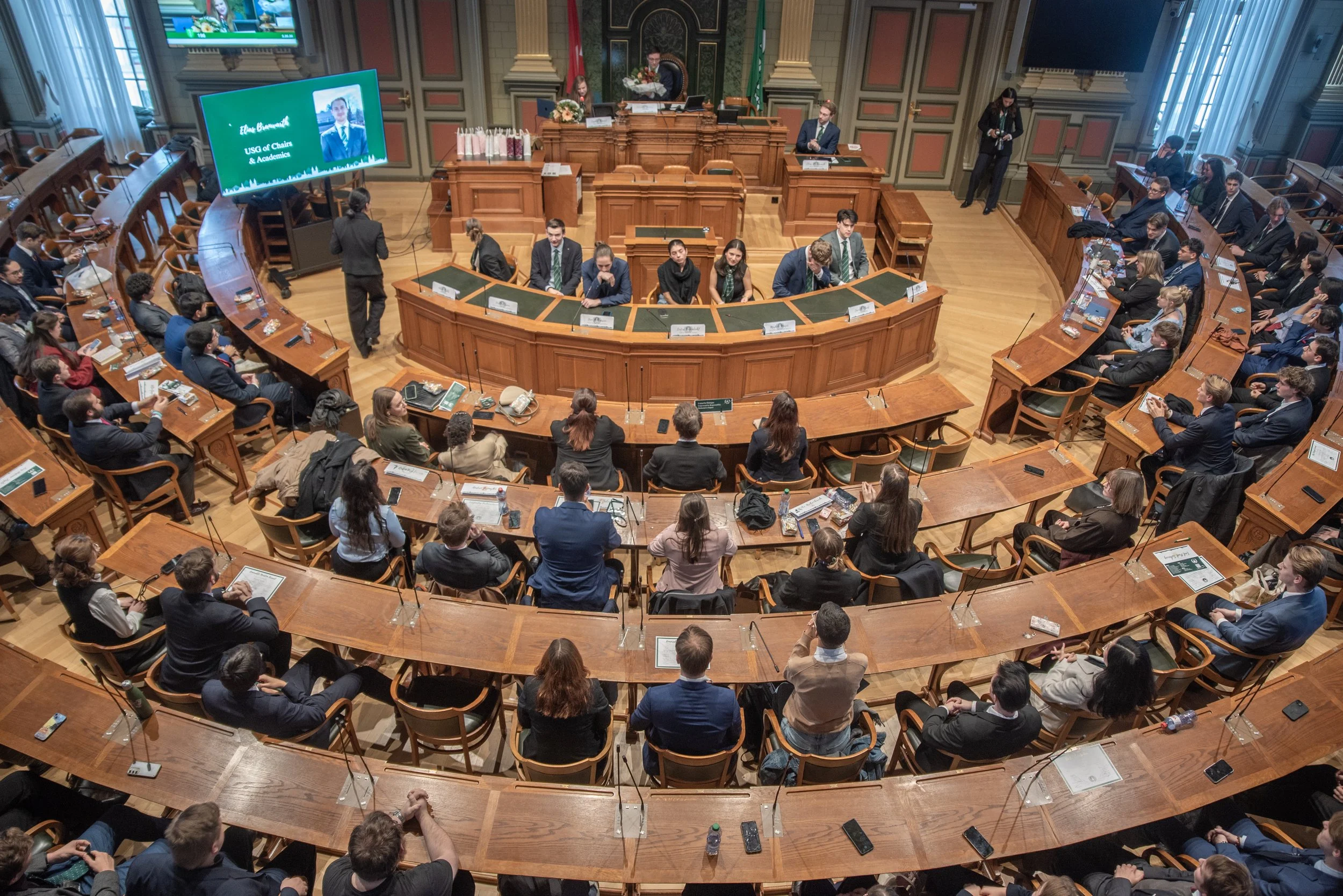 A large room filled with people sitting in a rounded, tiered wooden seating arrangement, attending a formal meeting or hearing. There is a podium at the front with a speaker, and a large screen displaying a presentation with the name 'Ellen Blankenship, USG of Chair & Academic'.