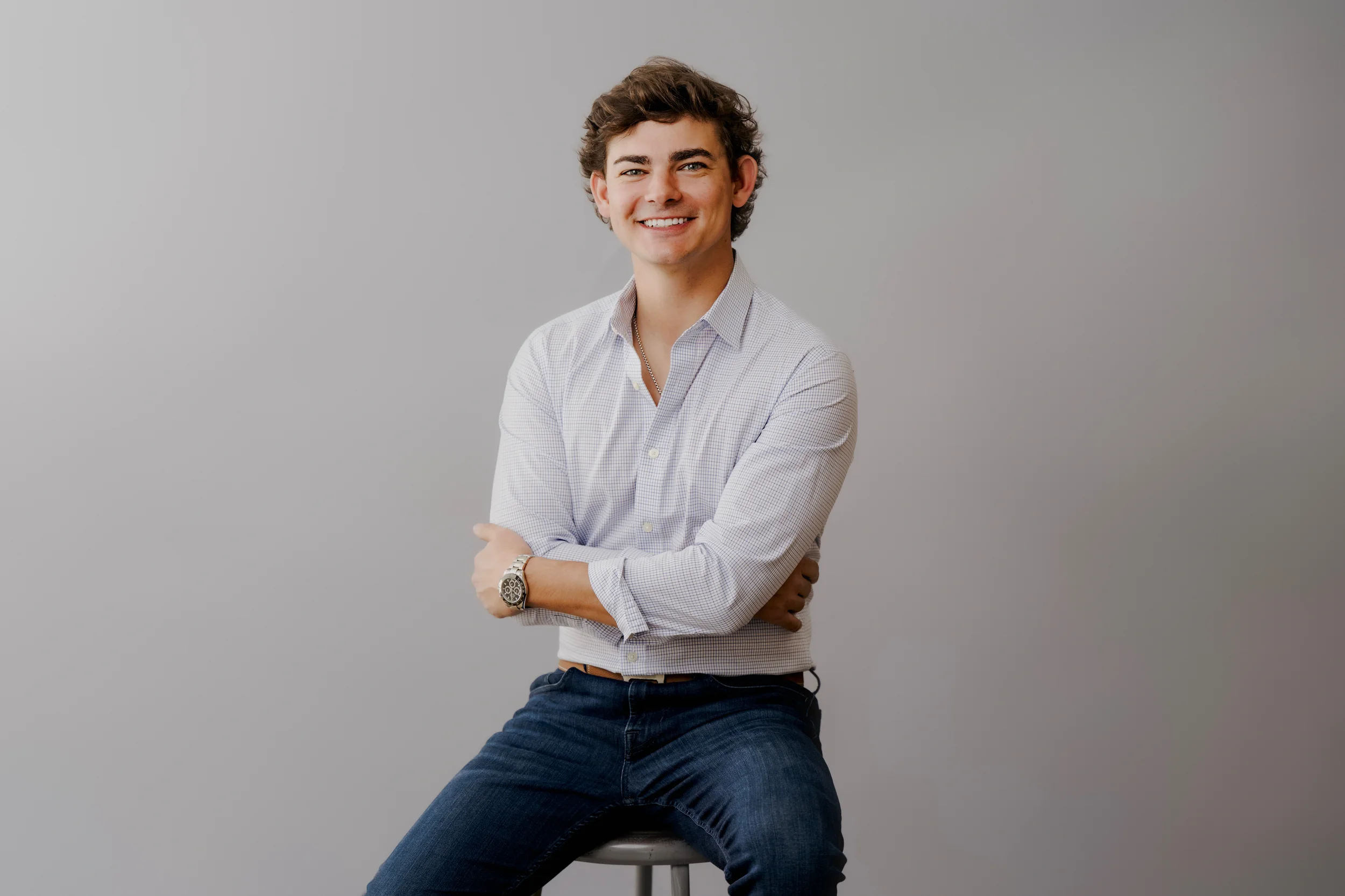 A young man with curly brown hair, wearing a light checkered button-up shirt, blue jeans, and a watch, sitting on a stool against a plain gray background, smiling at the camera.