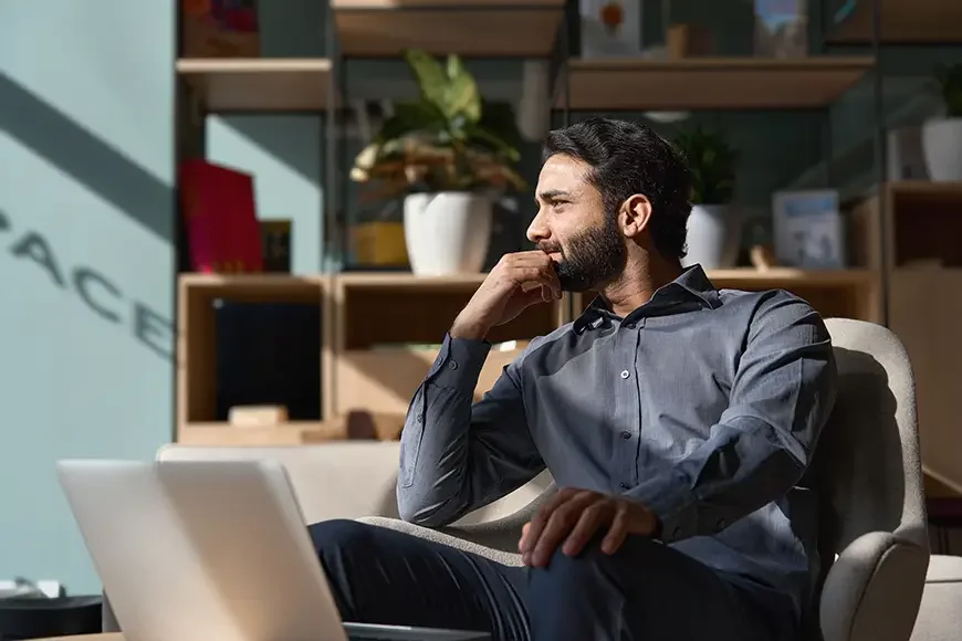 A man with dark hair and a beard, wearing a gray shirt, sitting on a beige chair in a modern office setting, looking thoughtfully at a laptop in front of him with shelves and potted plants in the background.