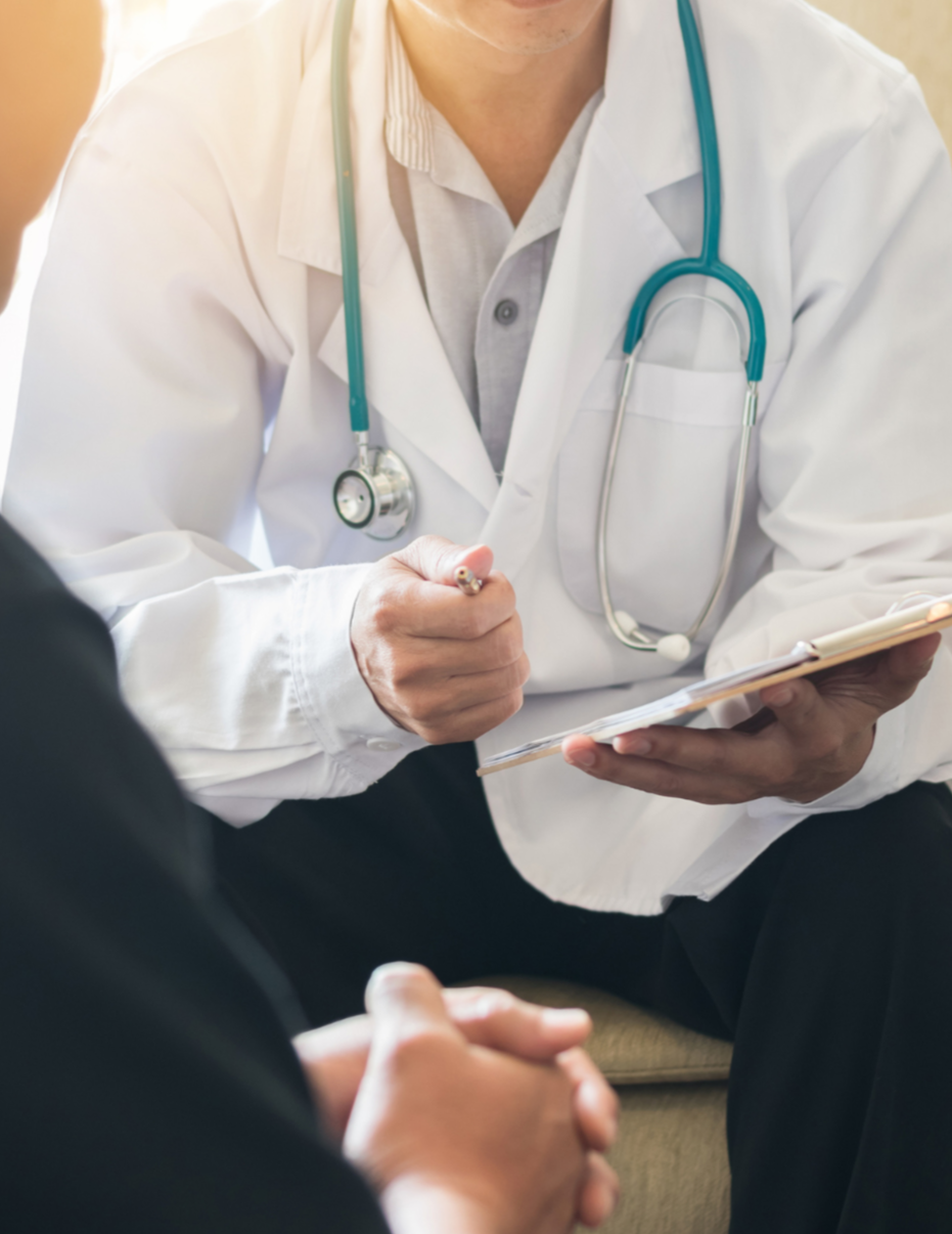 A male healthcare professional, likely a doctor, in a white coat with a stethoscope around their neck, holding a clipboard and talking to a patient.